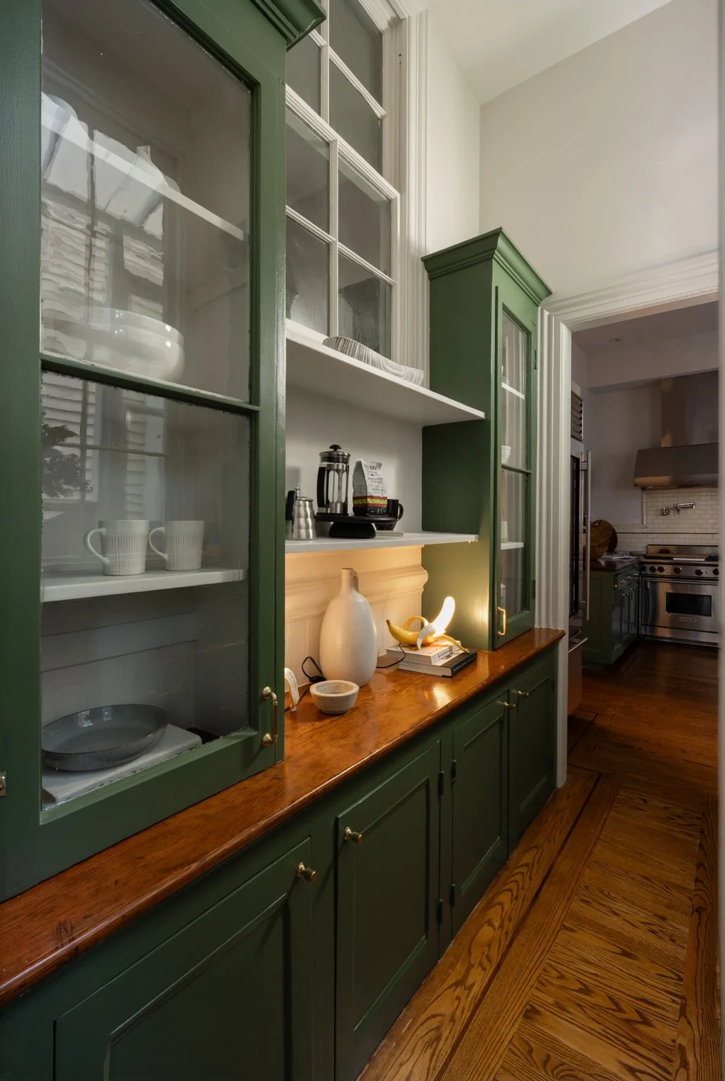 A kitchen with green cabinets and a wooden countertop, displaying bowls, cups, and decorative vessels, with a stove and range hood in the background.