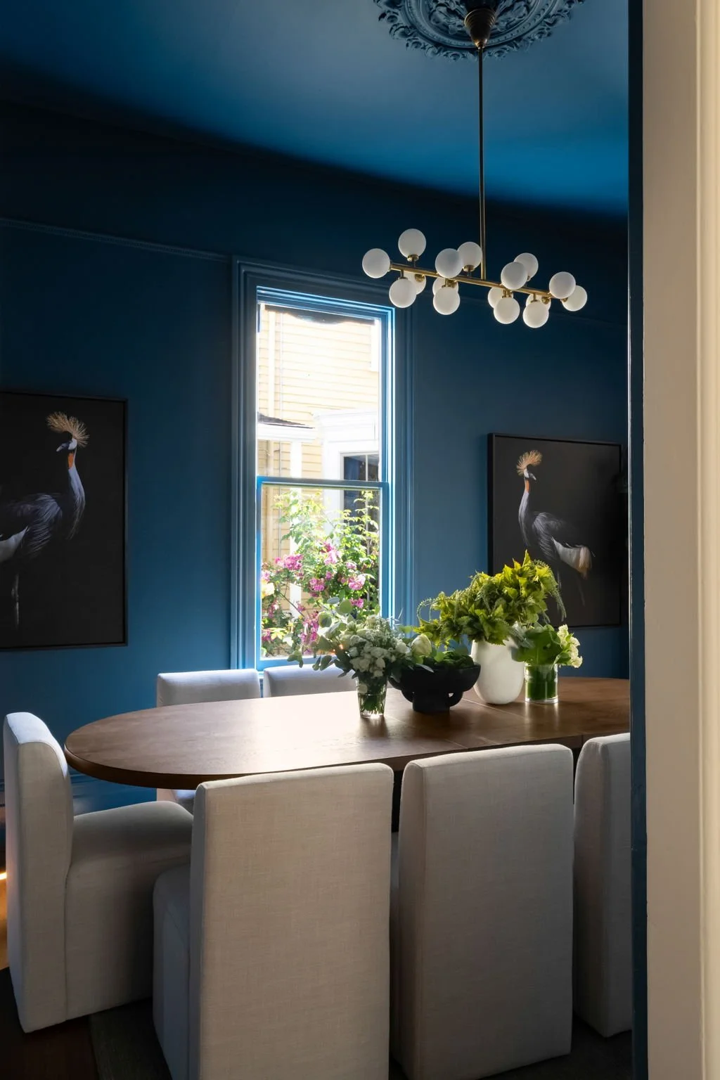 A dining room with dark blue walls, a wooden oval table surrounded by six white upholstered chairs, a window with sunlight and a view of blooming flowers, two black and white bird paintings, and a modern chandelier with spherical bulbs hanging from t
