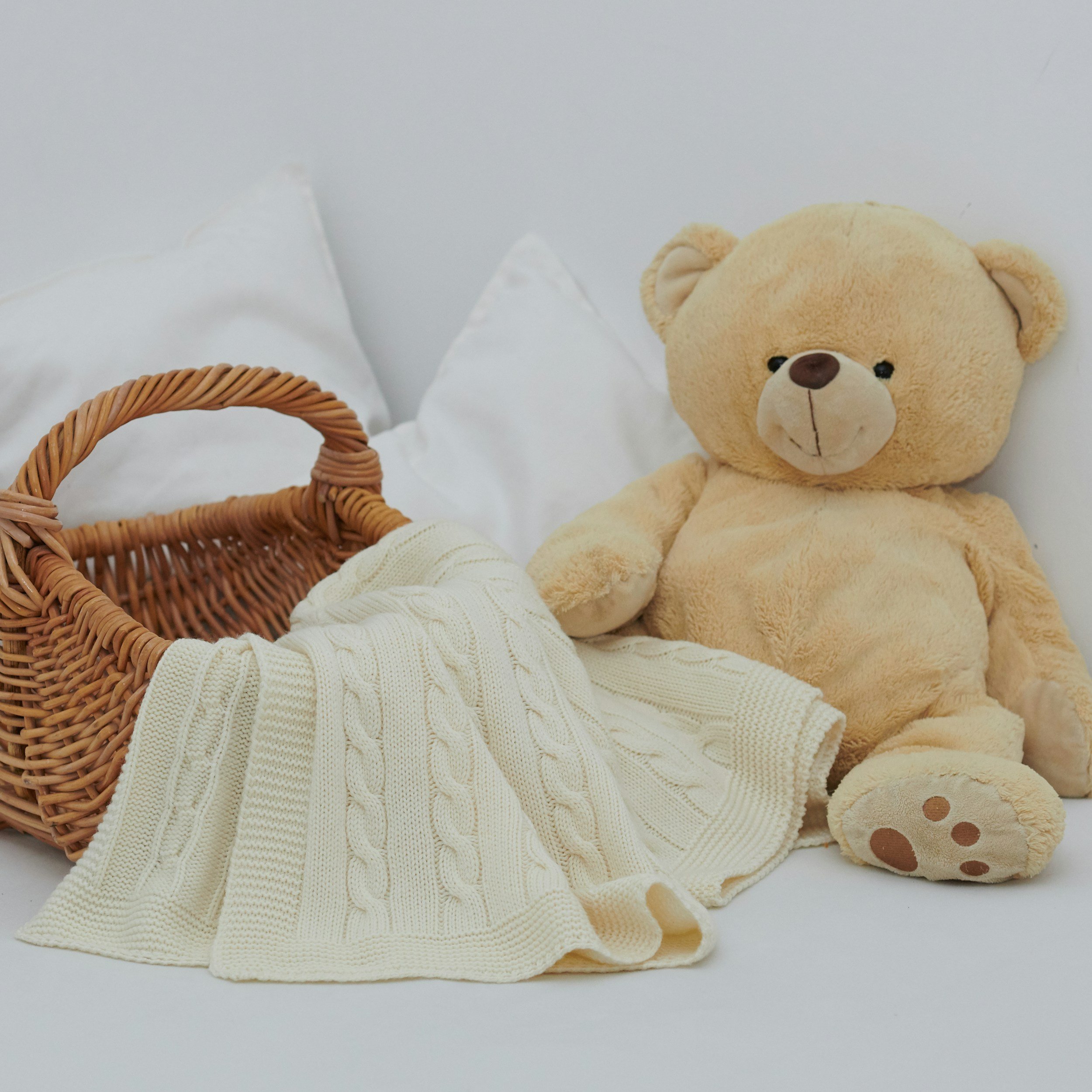 Stuffed teddy bear sitting on white bedsheets next to a wicker basket with a cream-colored blanket inside, with white pillows in the background.