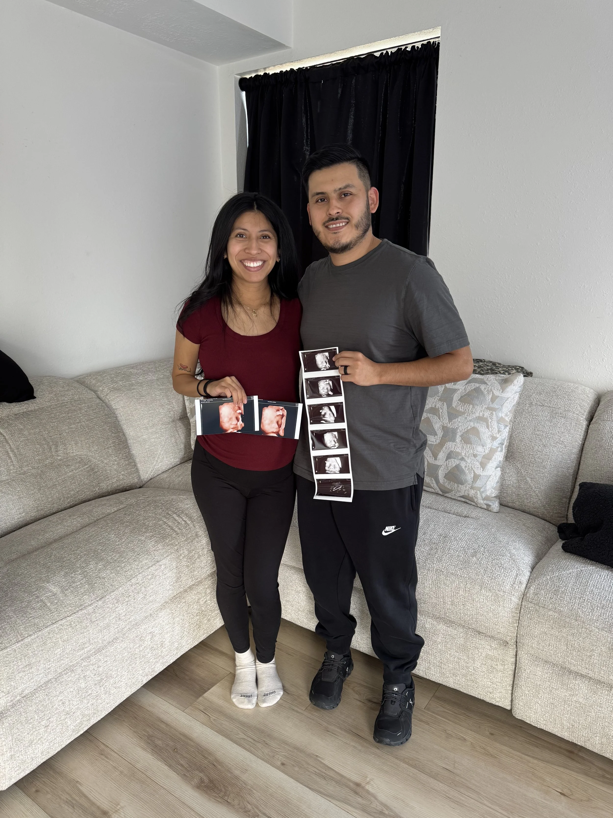 A smiling expectant couple sitting on a couch, proudly holding up a clear 3D ultrasound picture of their new baby.