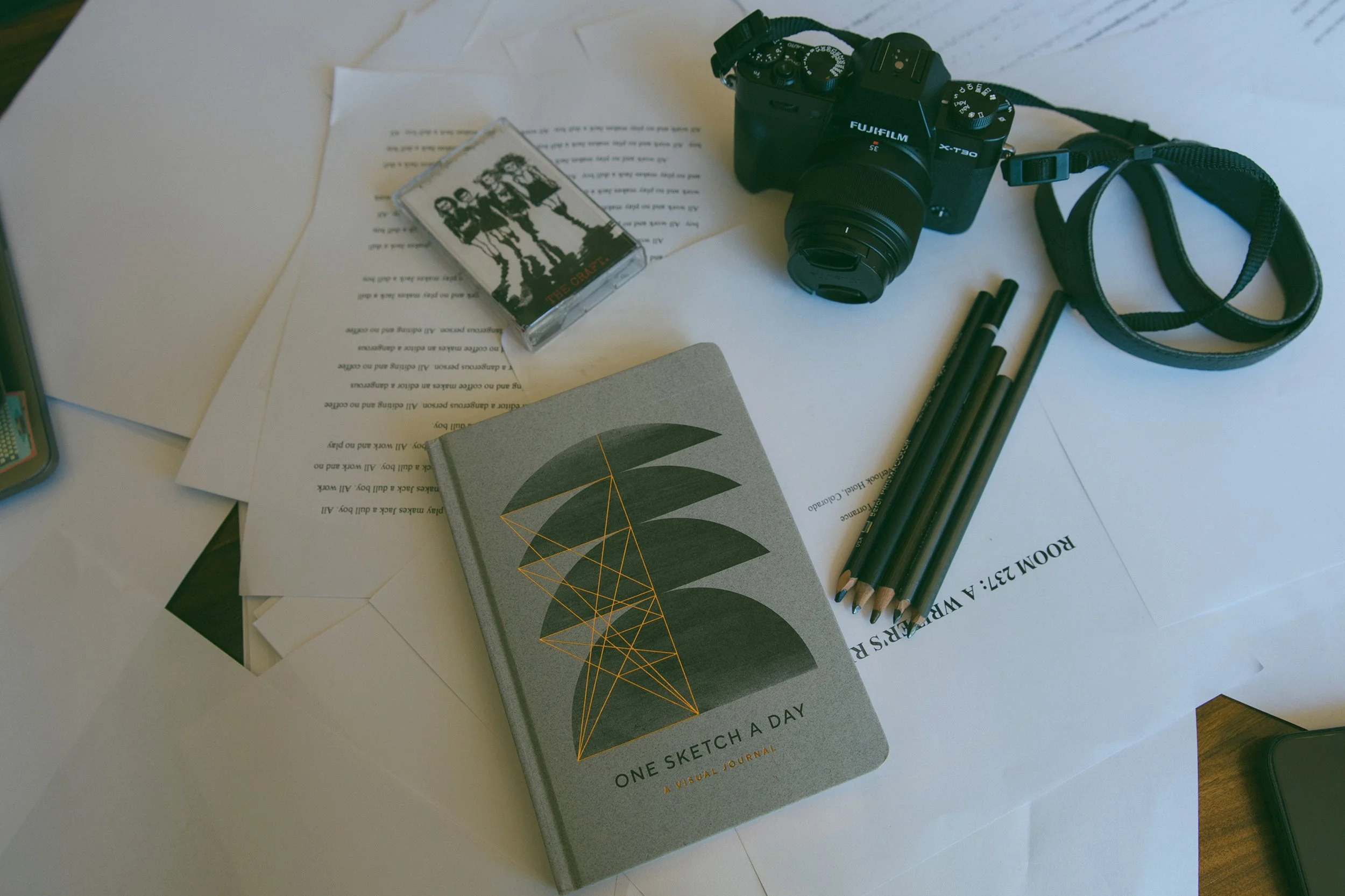 A mixtape, a Fujifilm camera, several pencils, and a sketchbook resting on the scattered pages of a manuscript