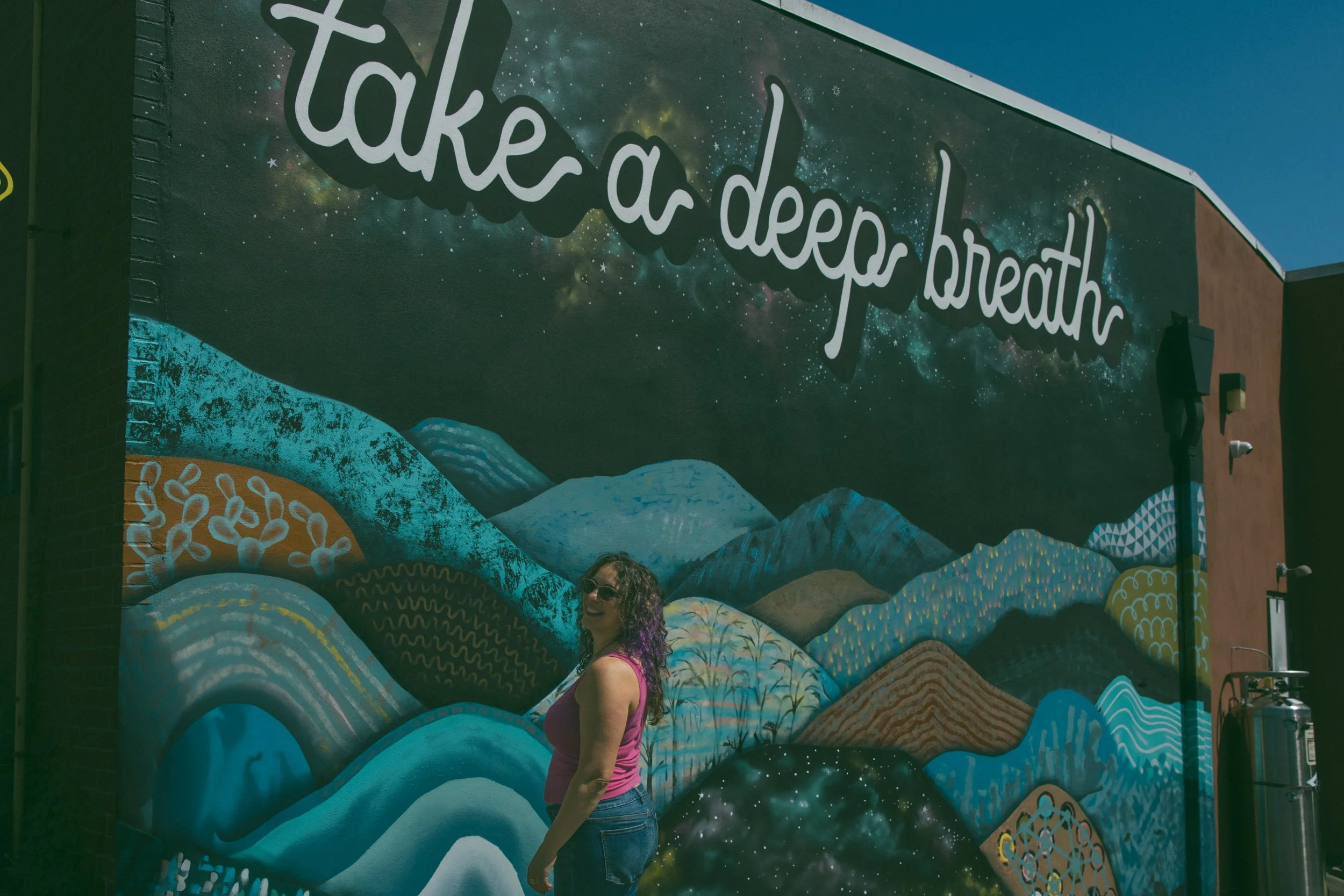 Samantha Pollack in a pink tank top against an outdoor mural that reads "take a deep breath"