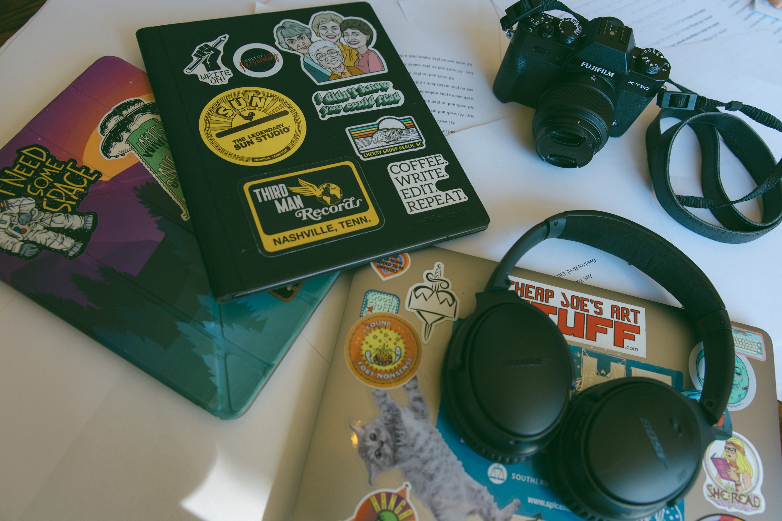 A cluttered work surface featuring a Fuji camera, over-ear headphones, and a laptop and tablet covered in stickers