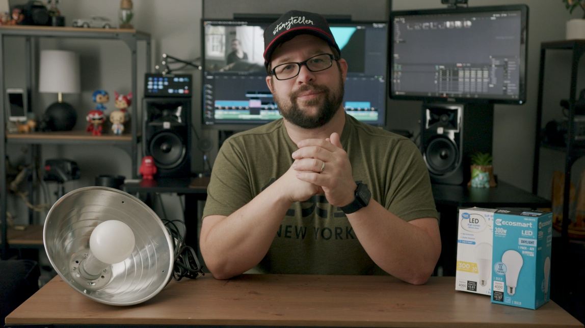 Man sitting at a desk in a room filled with computer monitors and audio equipment, holding hands together, with a large LED light bulb and a box of ecosmart LED bulb on the desk.