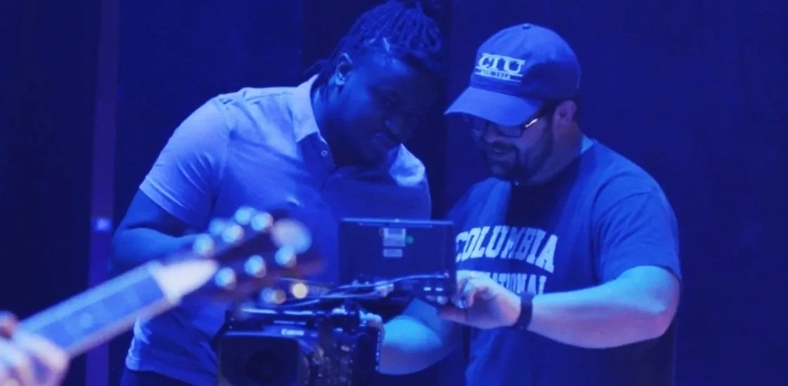 Two men looking at a tablet screen, with a guitar in the foreground, in a dimly lit blue room.