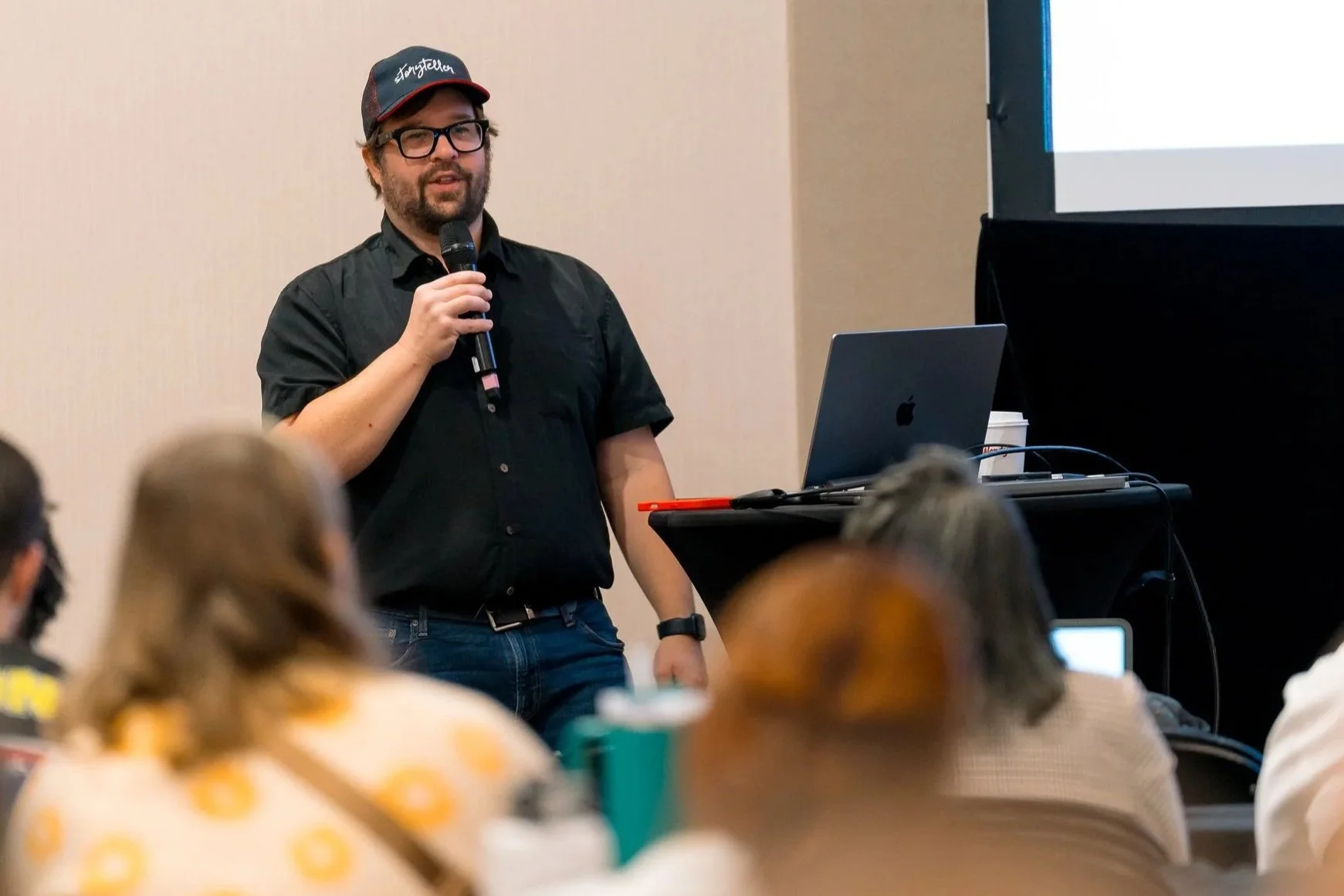 A man with glasses and a beard wearing a black shirt and a baseball cap with the word 'Jangstler' in white text, speaking into a microphone during a presentation. He is standing next to a laptop on a table in a conference room with people seated and listening.