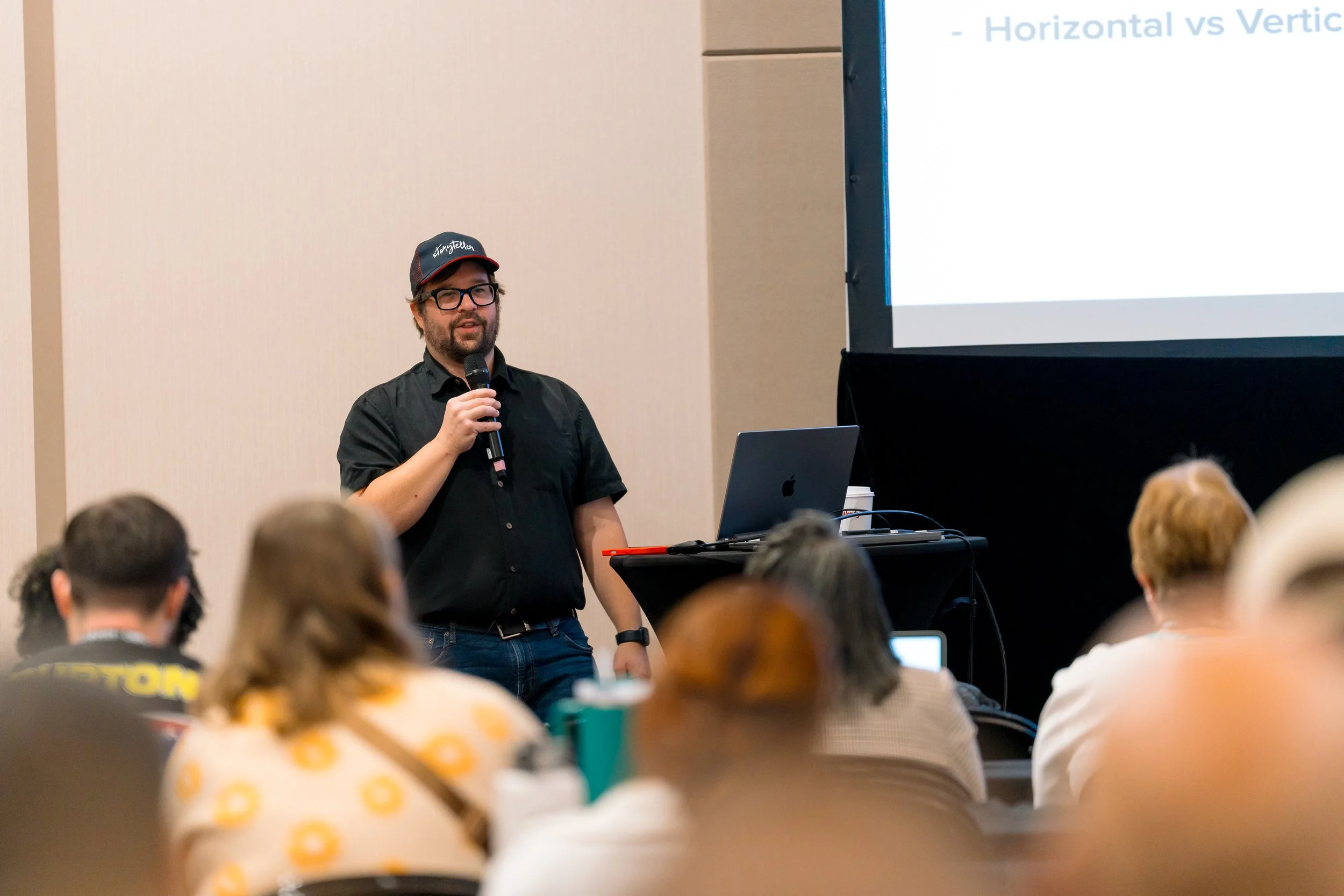 A man wearing glasses, a black shirt, and a cap is giving a presentation in a conference room, holding a microphone. There is a laptop on a table next to him and a large screen displaying a slide titled 'Horizontal vs Vertical.' Several attendees are seated, watching the presentation.