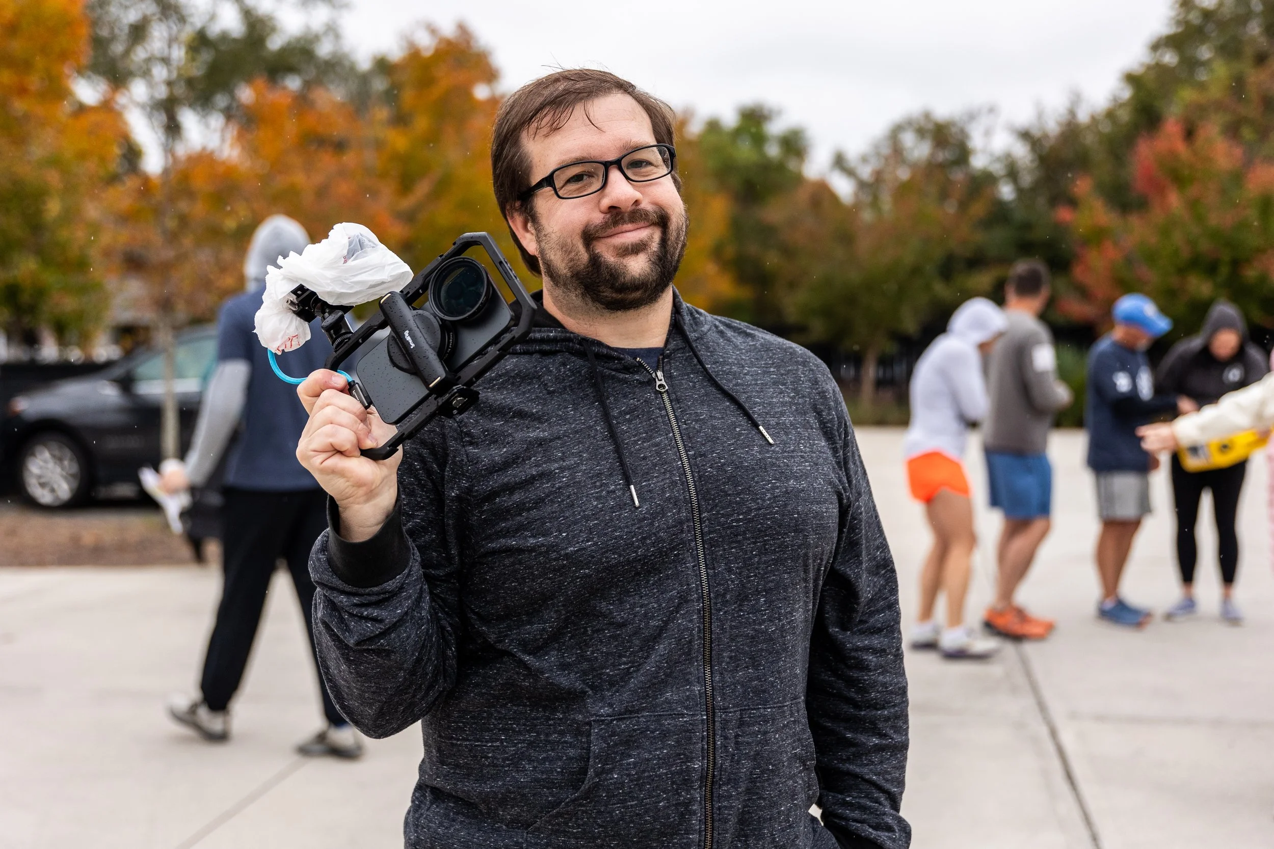 A man with glasses and a beard in a black sweatshirt holding a camera with a crisis mask attached, standing outdoors with trees showing fall foliage in the background, and others in casual attire in the distance.