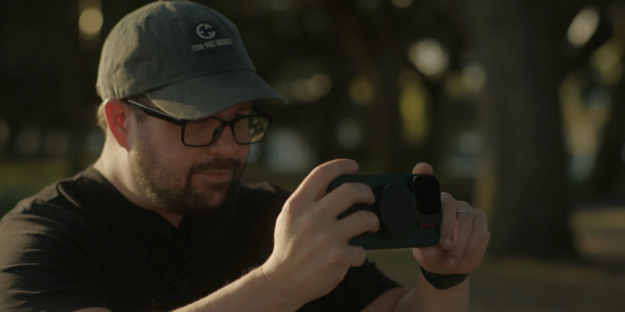 Man with glasses and a gray baseball cap taking a photo or video with a smartphone outdoors in park in late afternoon