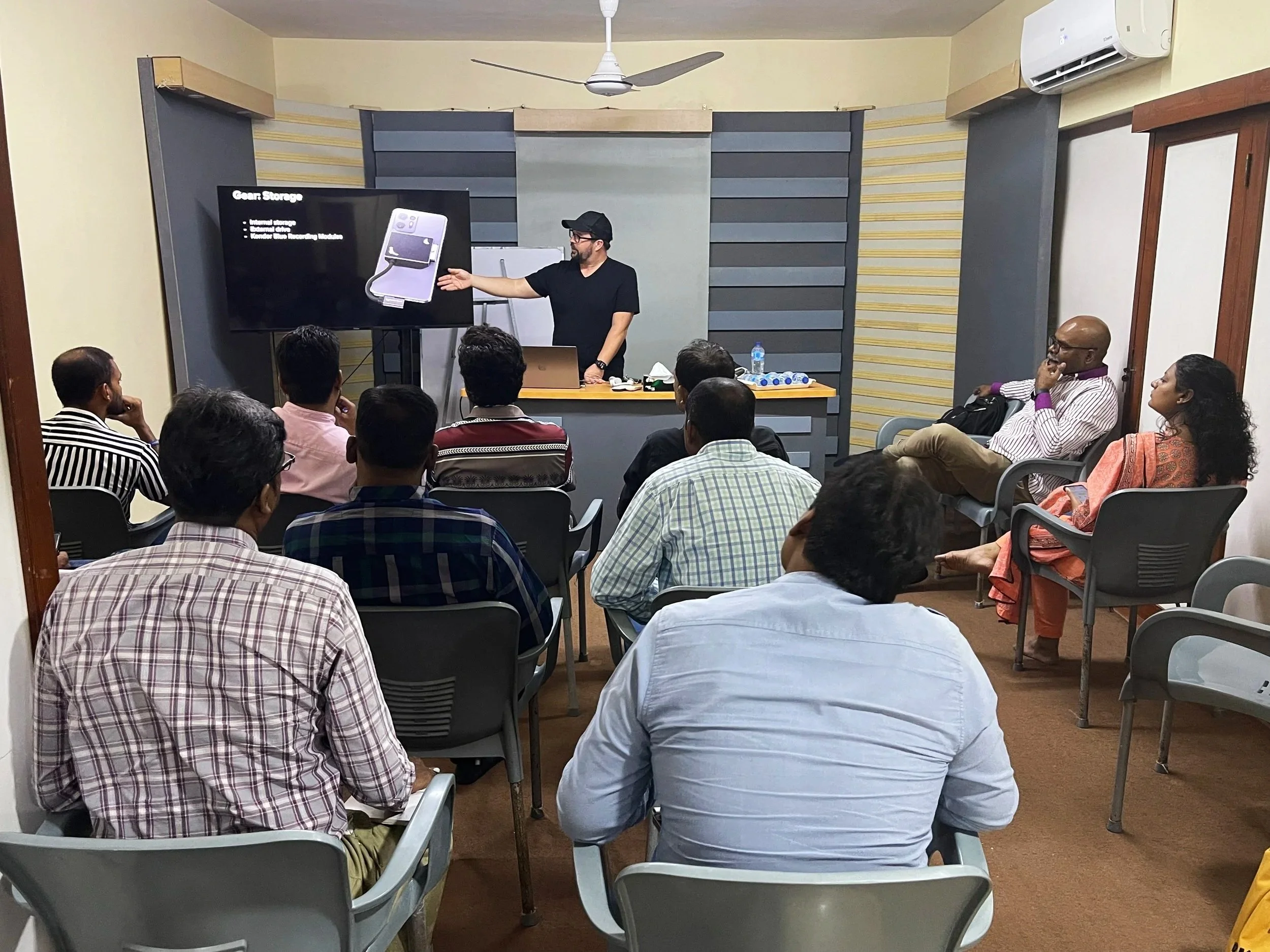 A man in black T-shirt and cap is giving a presentation to an audience in a classroom or conference room. The presenter is pointing at a large screen displaying a slide about gear storage with an image of a device or equipment. The audience, consisting of men and women, sit on chairs facing the screen, attentively watching the presentation. The room has beige walls, a ceiling fan, and an air conditioning unit.