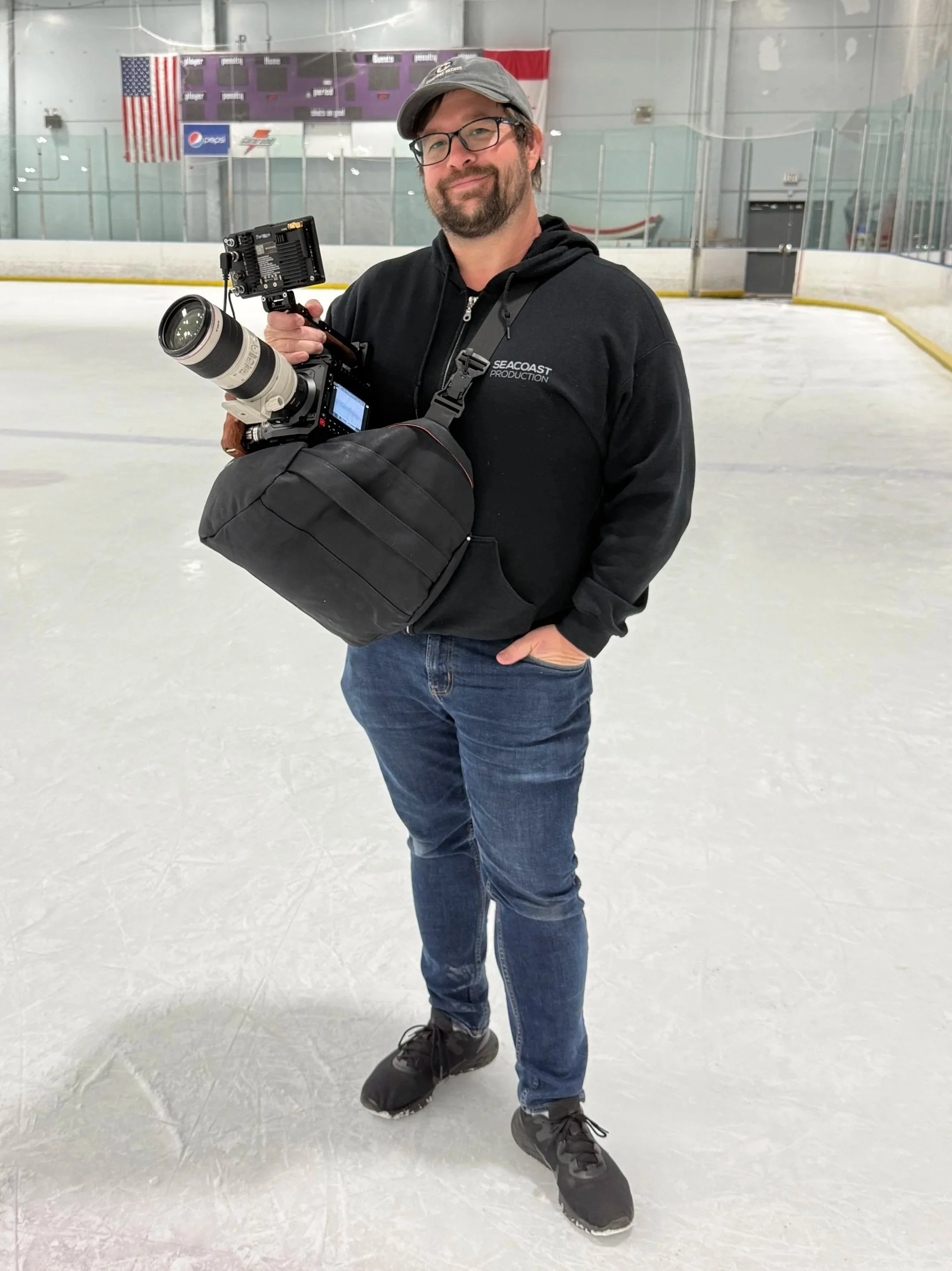 A man standing on an ice rink holding a professional camera with a large lens, wearing a black hoodie, jeans, a gray cap, and glasses, with a camera bag strapped to his shoulder.