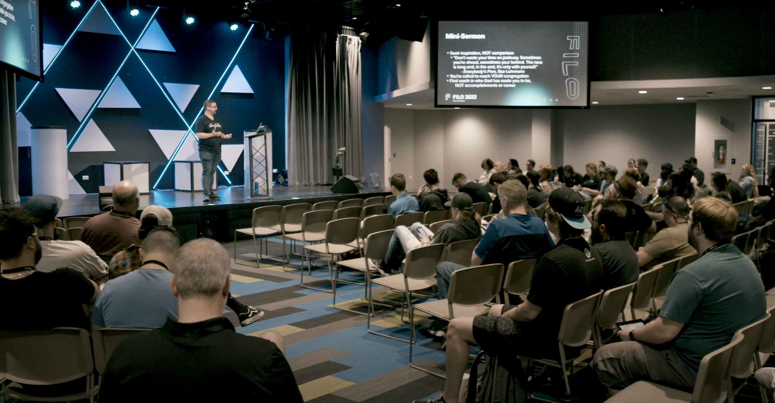 A speaker on stage at a conference giving a presentation about a mini-sermon with an audience seated in chairs watching the presentation.