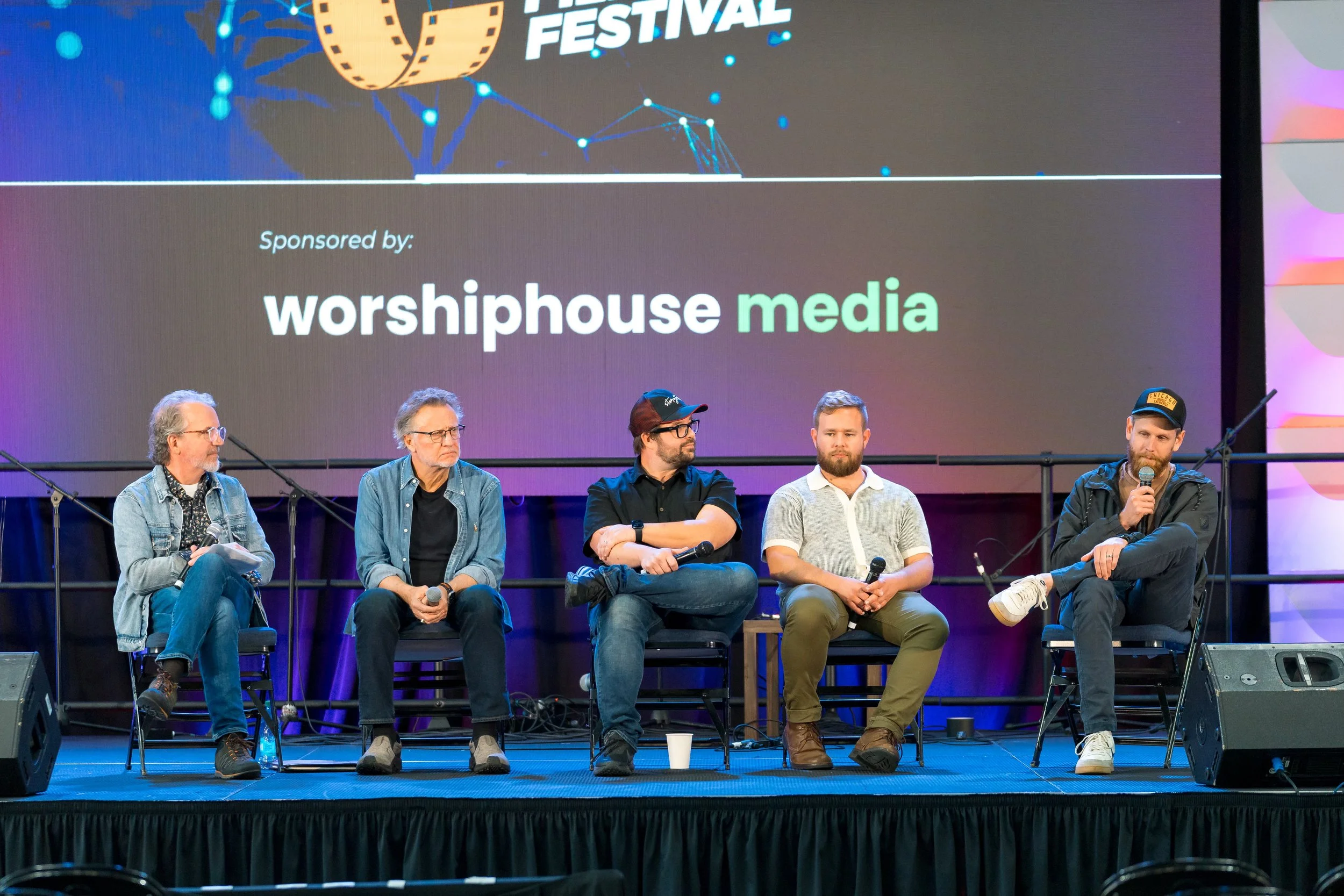 A panel of five men sitting on a stage during a conference, with a large screen behind them displaying event sponsorship and media logos.