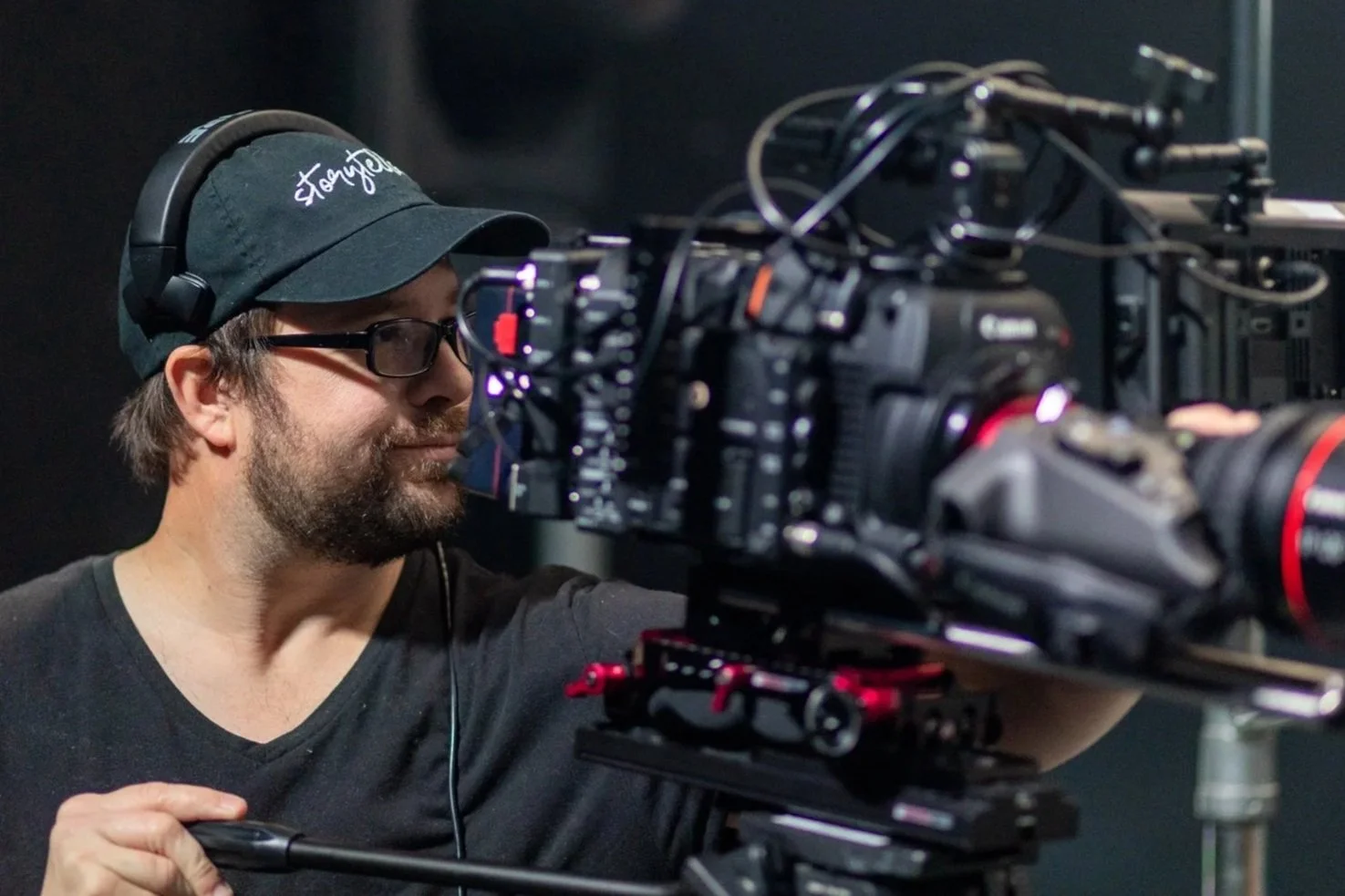 A camera operator wearing glasses, a black baseball cap, and headphones films with a professional video camera.
