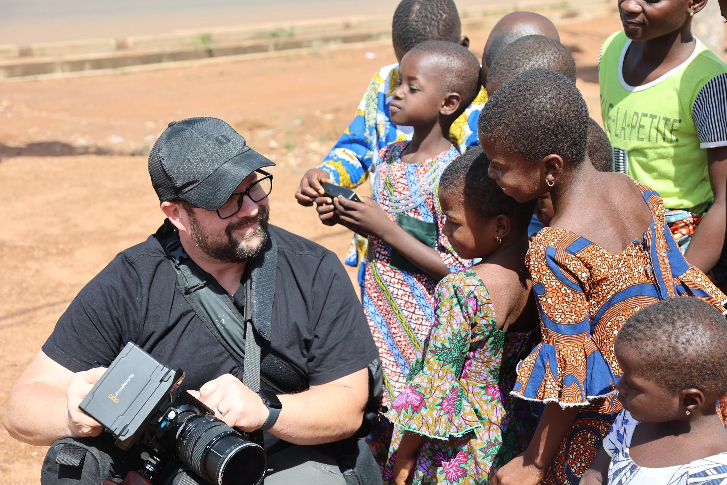 A man with glasses and a black baseball cap, holding a camera, is smiling and surrounded by smiling children in colorful clothing during daytime outdoors.