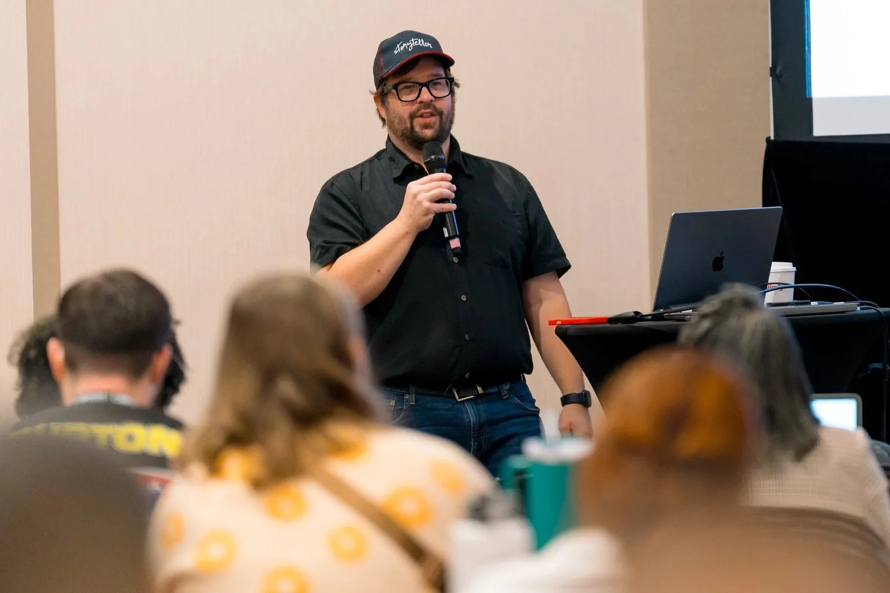 A man with glasses and a beard wearing a black shirt and baseball cap gives a presentation with a microphone in front of an audience, with a laptop on a stand beside him.