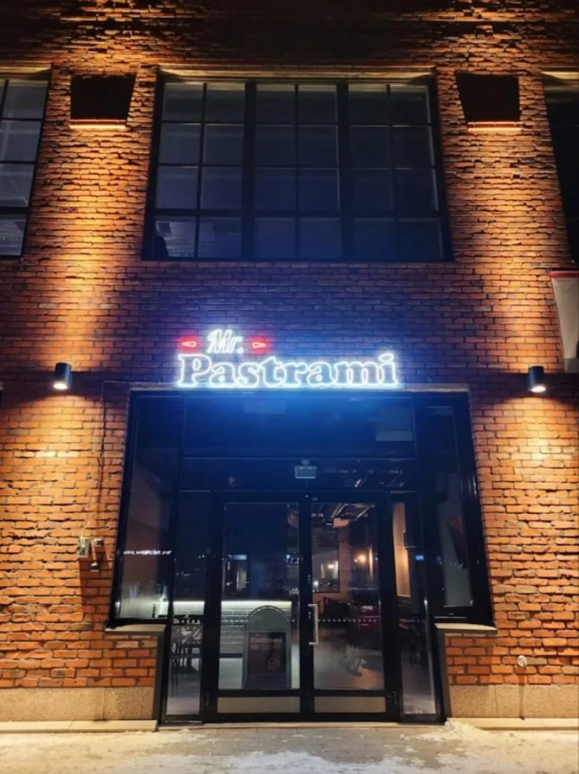 Ravintolan tai kahvilan sisäänkäynti, jossa neonkyltti lukee "Mr. Pastrami". Rakennus on tiilistä, ja ikkunoissa näkyy sisätiloja.