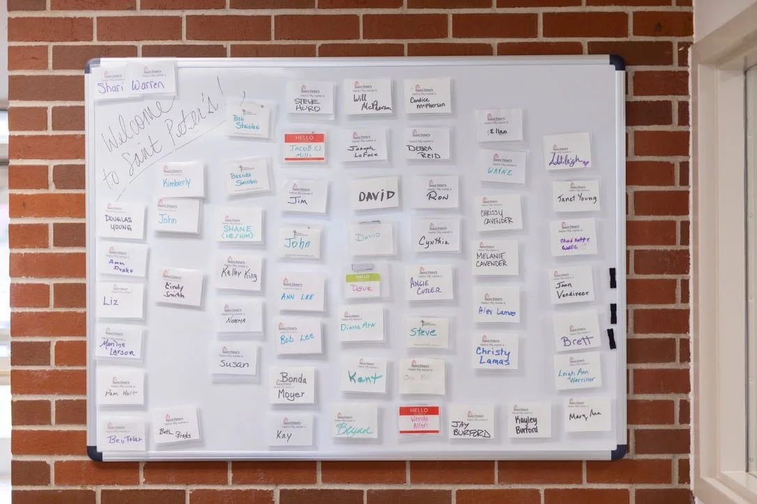 Whiteboard with multiple name tags, welcome message, and notes, mounted on a brick wall.