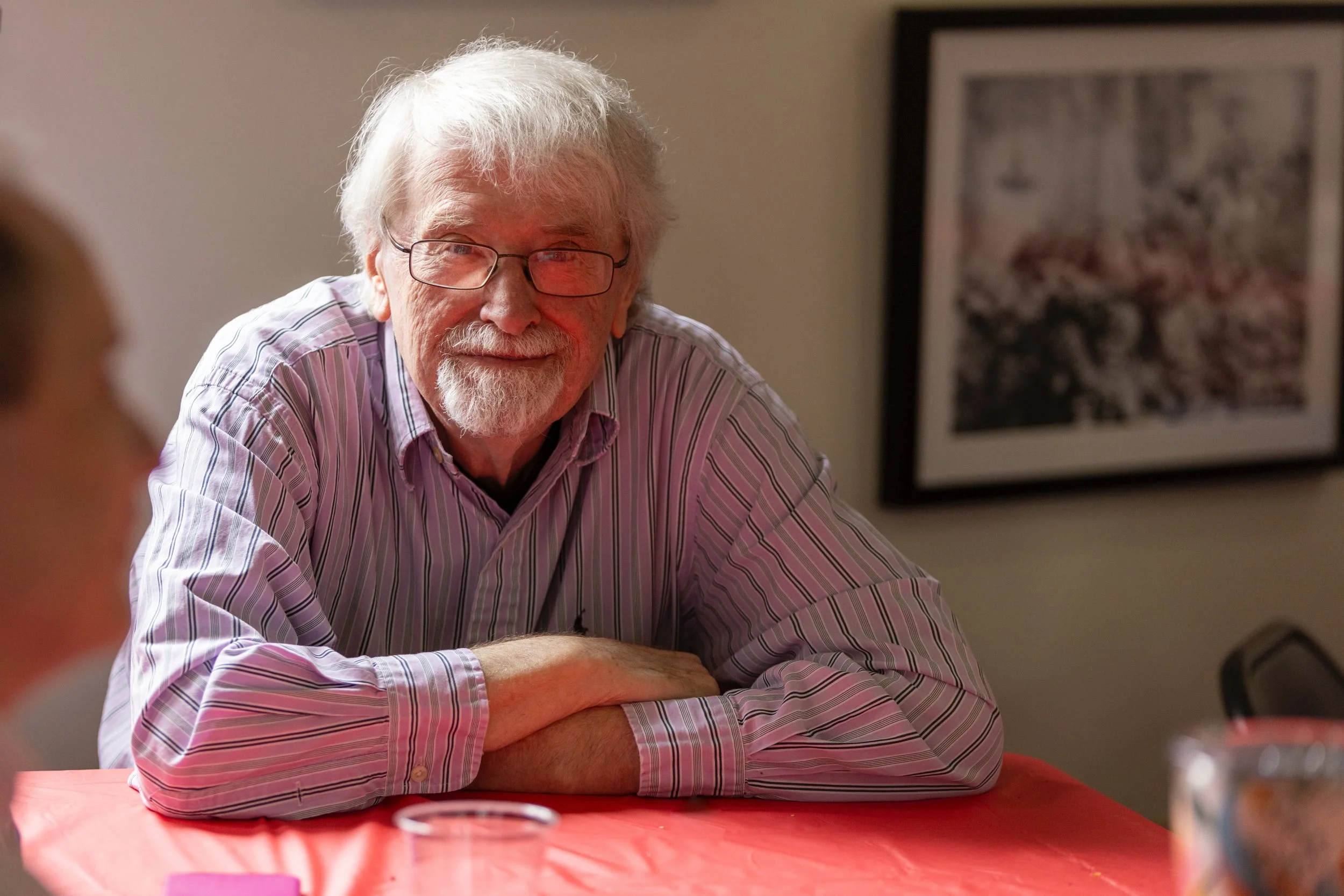 An elderly man with gray hair, glasses, and a beard, smiling, sitting at a table with a red tablecloth.