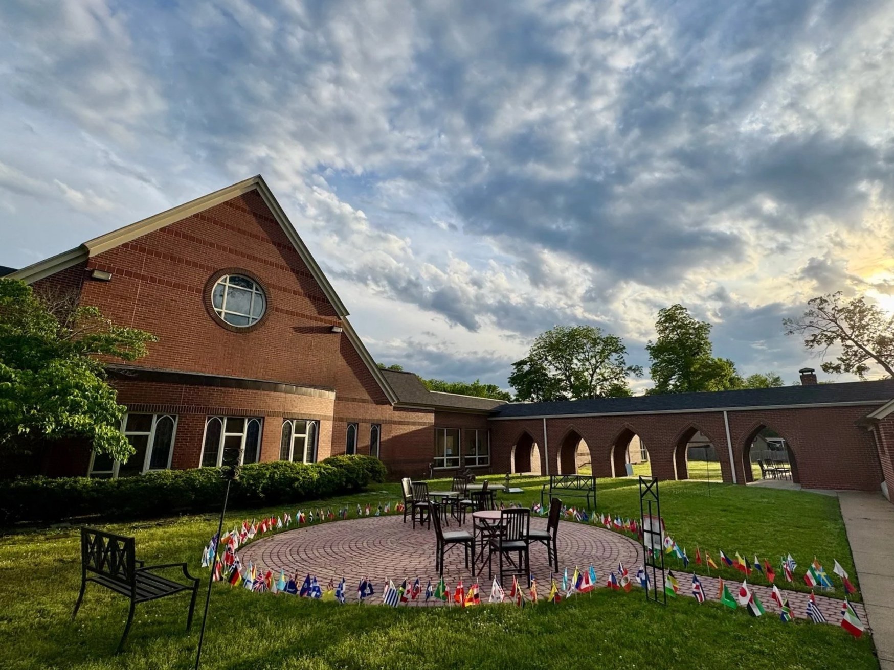 Red brick church with a tall steeple, surrounded by green trees, benches, and a grassy area with a brick labyrinth in front of the church.