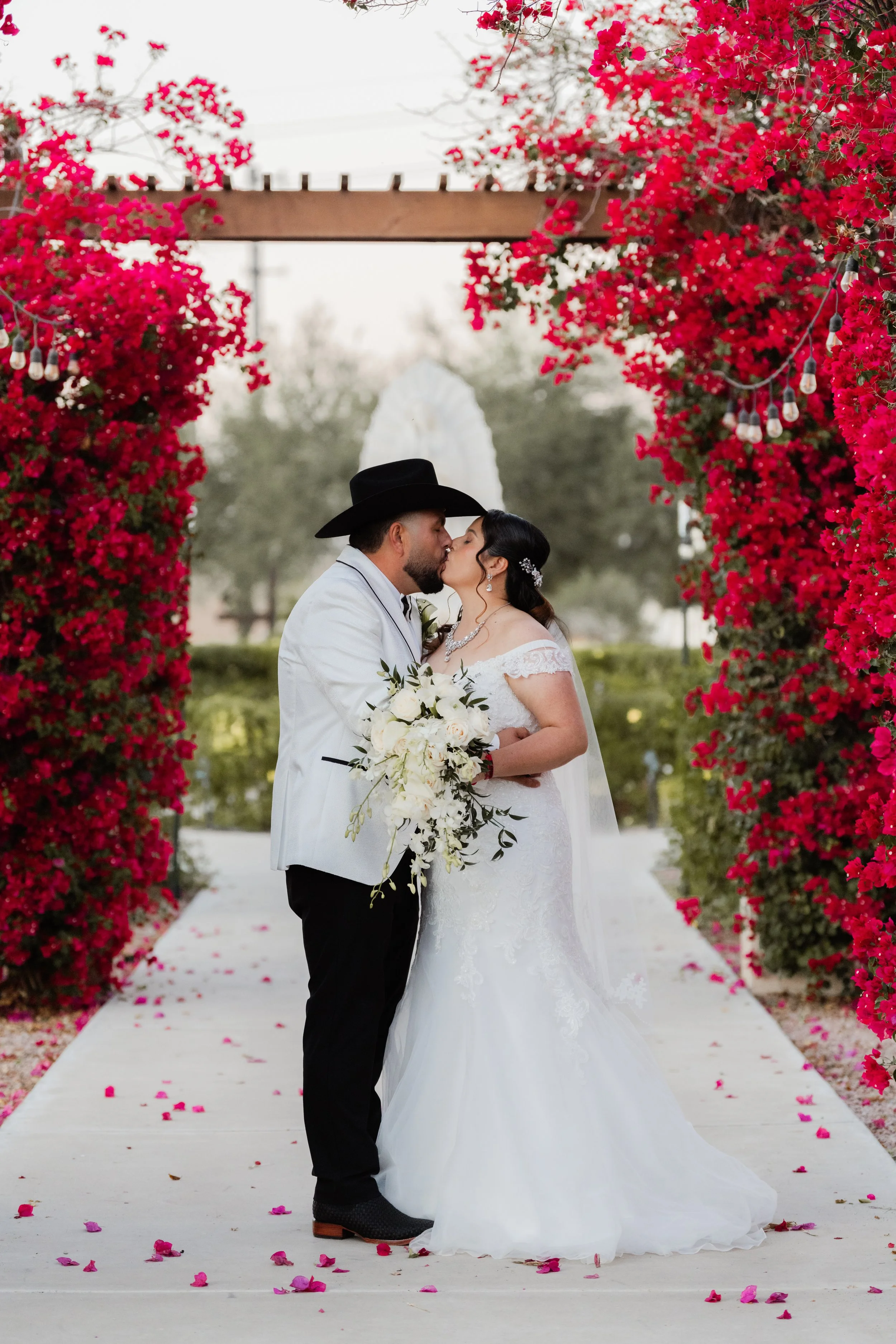 A bride and groom sharing a kiss during their wedding ceremony, surrounded by vibrant pink flowers and decorative lights.