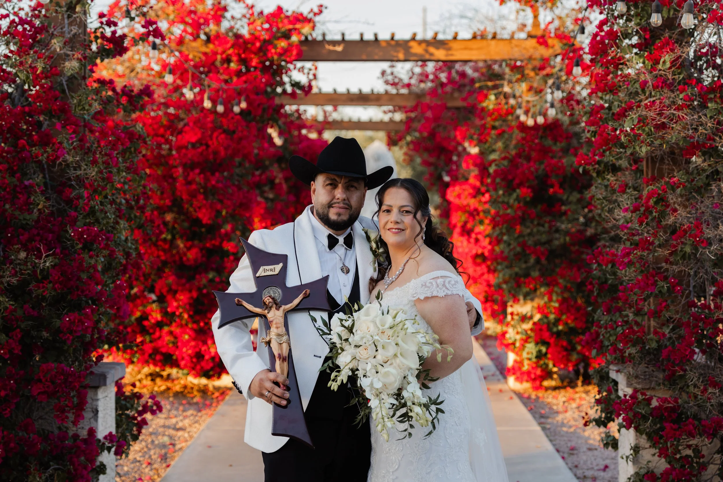 Wedding couple standing on a pathway surrounded by vibrant pink and red bougainvillea flowers, with a wooden pergola overhead. The groom is wearing a white tuxedo jacket, black bow tie, and a cowboy hat, holding a crucifix. The bride is in a white lace wedding dress, holding a bouquet of white flowers, smiling at the camera.
