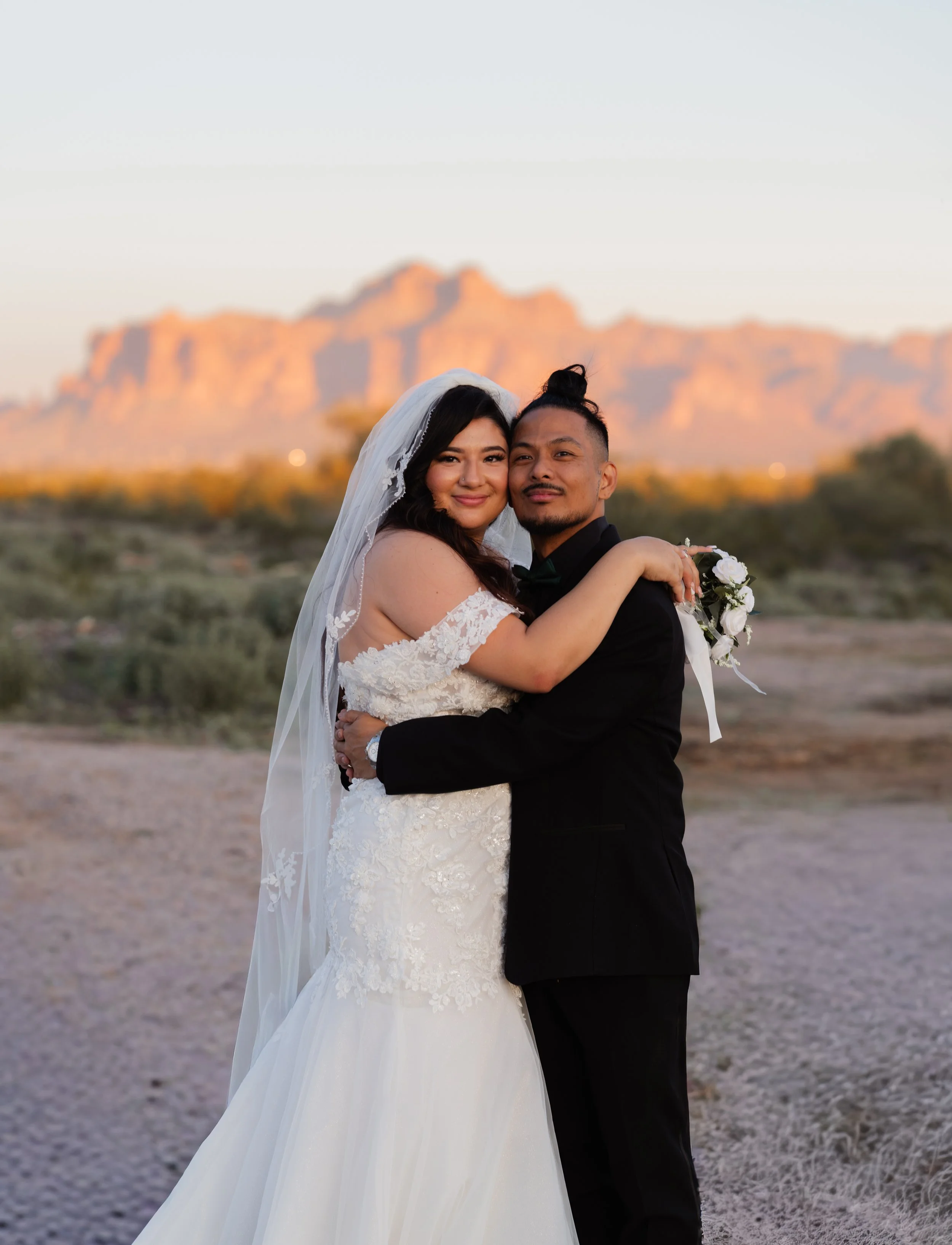 A newlywed couple, a woman in a white lace wedding dress and a veil, and a man in a black suit, are embracing outdoors at sunset, with a mountain range in the background.