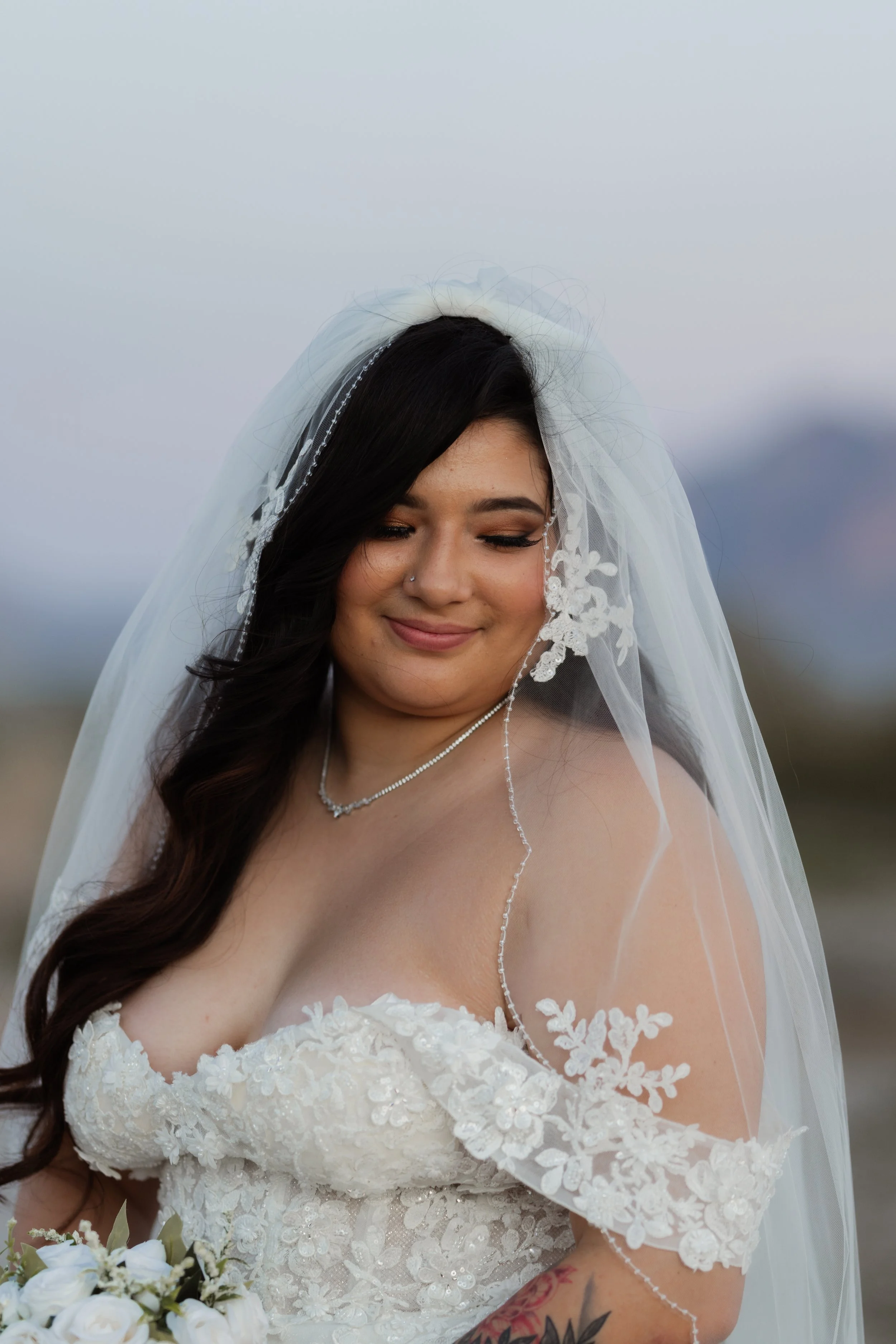 A bride with long dark hair, wearing a lace off-the-shoulder wedding dress and a veil, holding a bouquet of white roses, smiling softly outdoors.