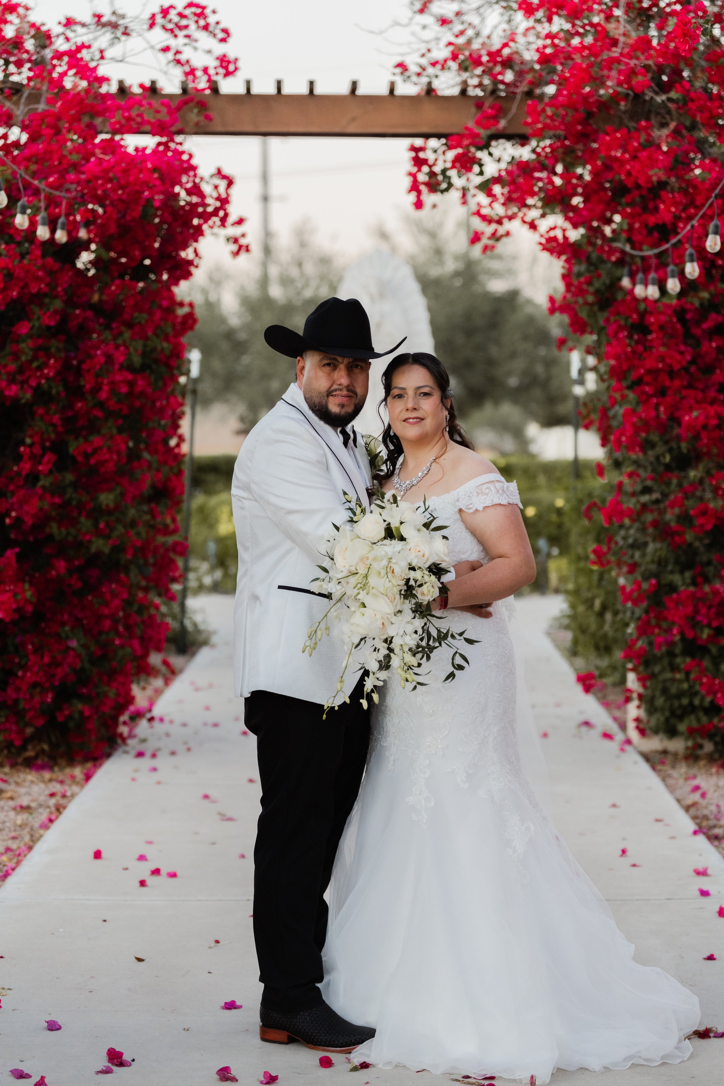A bride and groom standing under a floral arch at their wedding, with pink bougainvillea flowers and a white pathway decorated with rose petals.