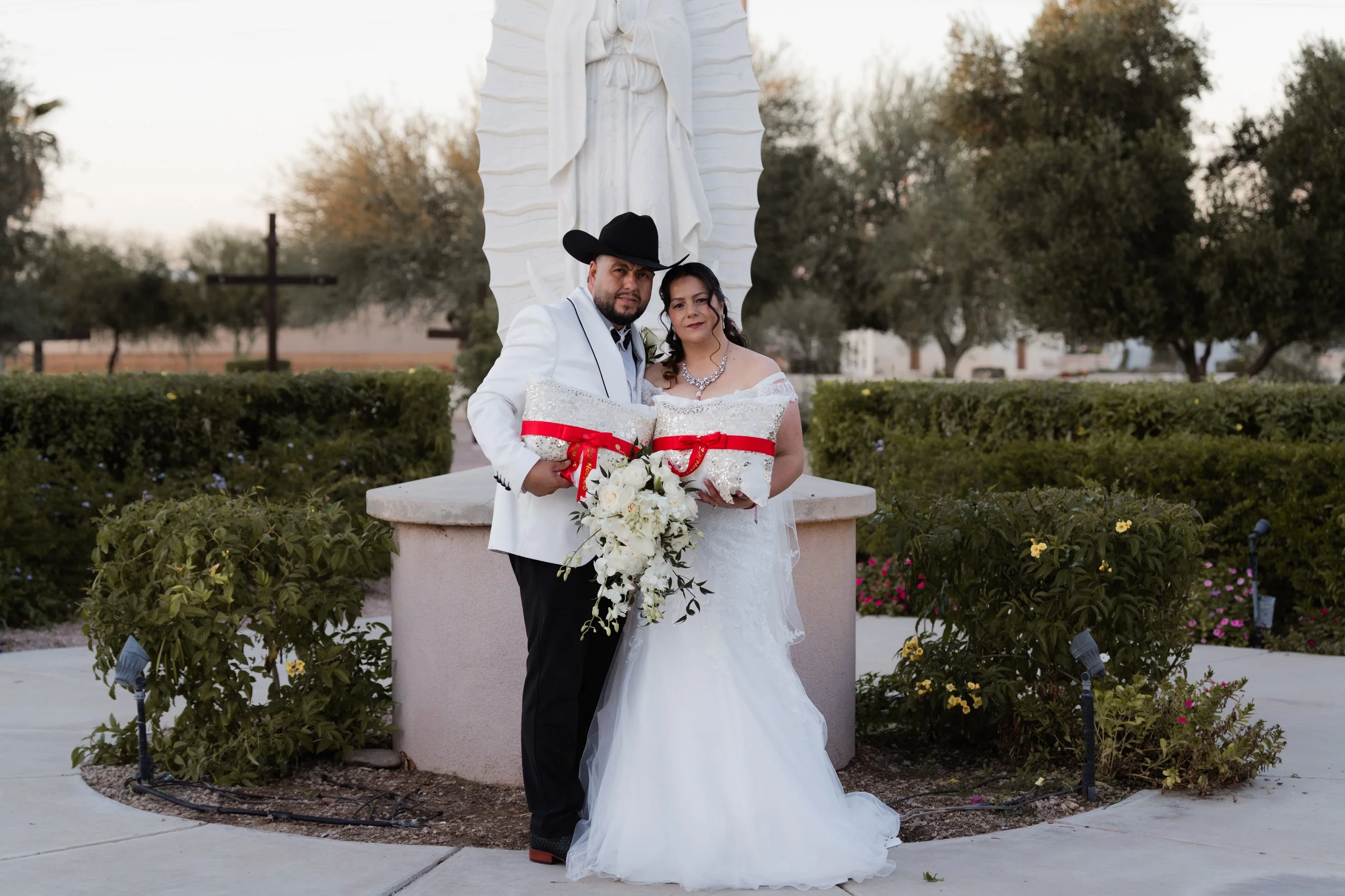A newlywed couple posing outdoors beside a religious statue, with the groom in a white tuxedo and cowboy hat holding a pillow with a red ribbon, and the bride in a white wedding gown holding a bouquet of white flowers.