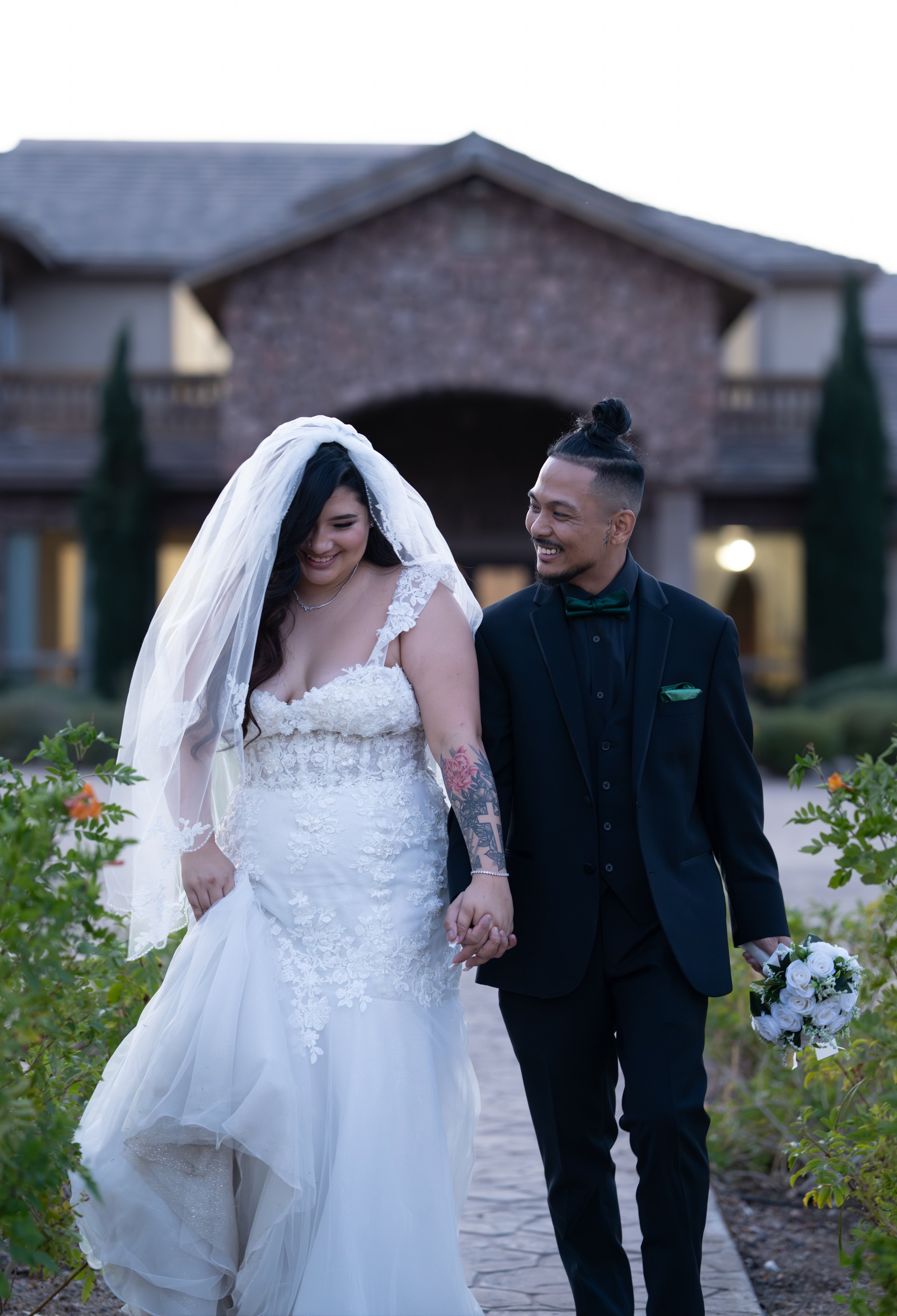 A newlywed couple walking hand in hand outside a house, smiling and dressed in wedding attire, with the bride wearing a veil and gown, and the groom in a black suit with a green bow tie, holding a bouquet of white roses.