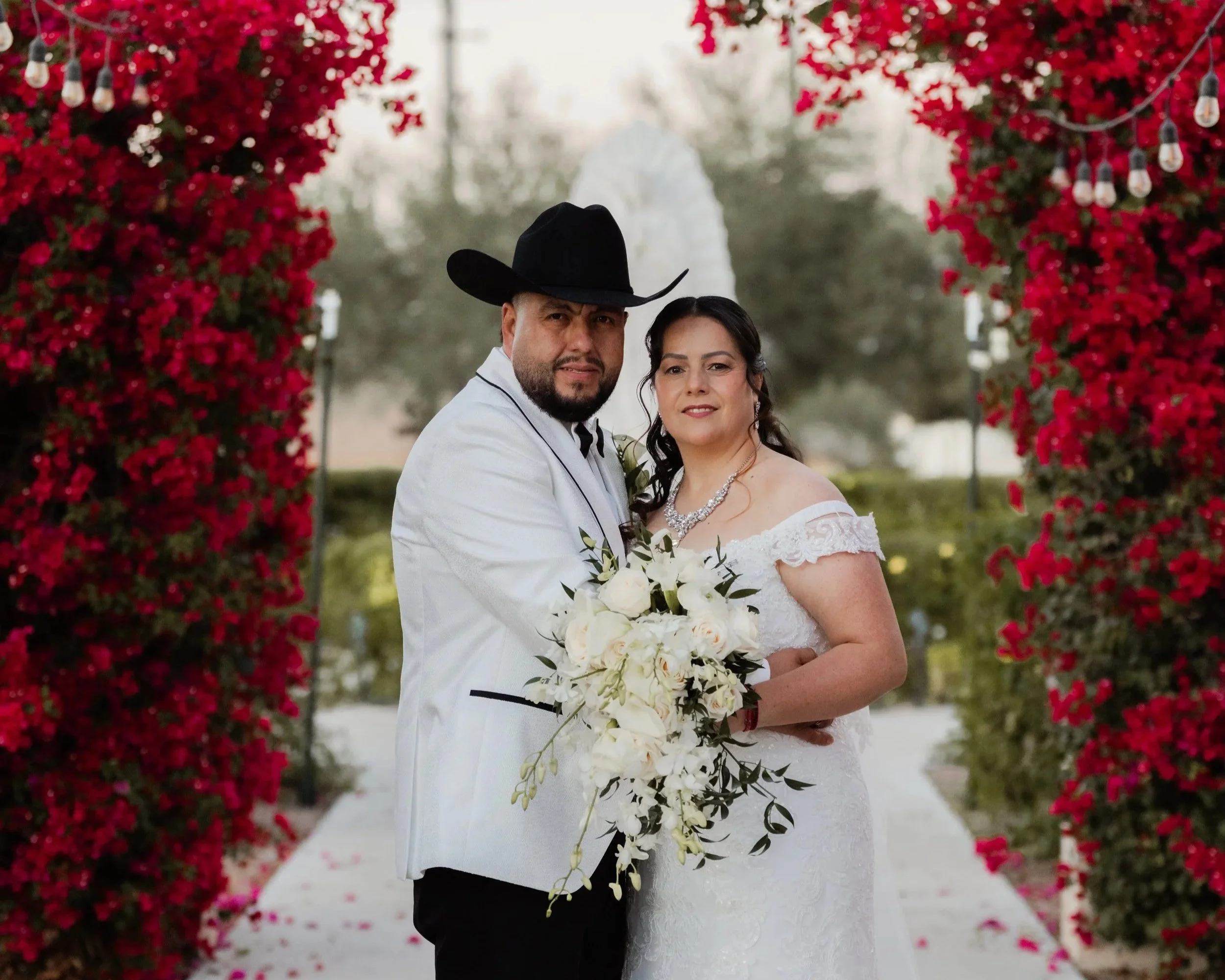 A newlywed couple posing together outdoors, framed by a floral arch with pink flowers and string lights, with a background of trees and sky.