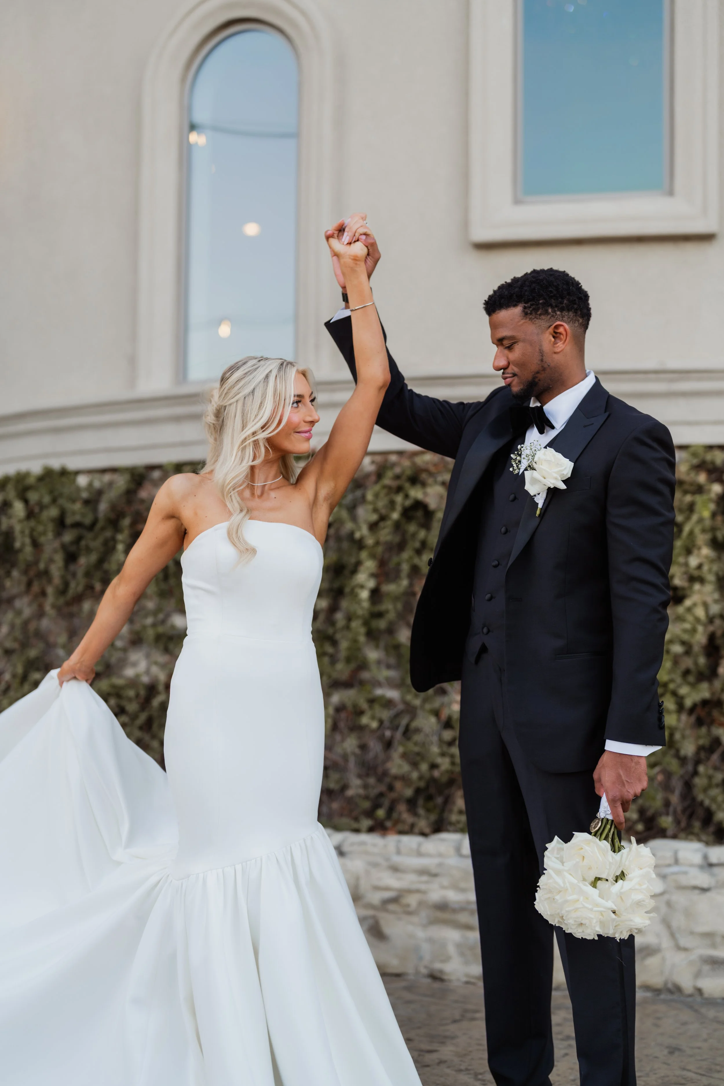 A bride and groom dancing outdoors on a wedding day, holding hands up in a gentle dance pose. The bride is in a strapless white wedding dress, and the groom is in a black tuxedo with a white boutonniere and a bouquet of white flowers in his hand. There are arched windows and a building in the background.