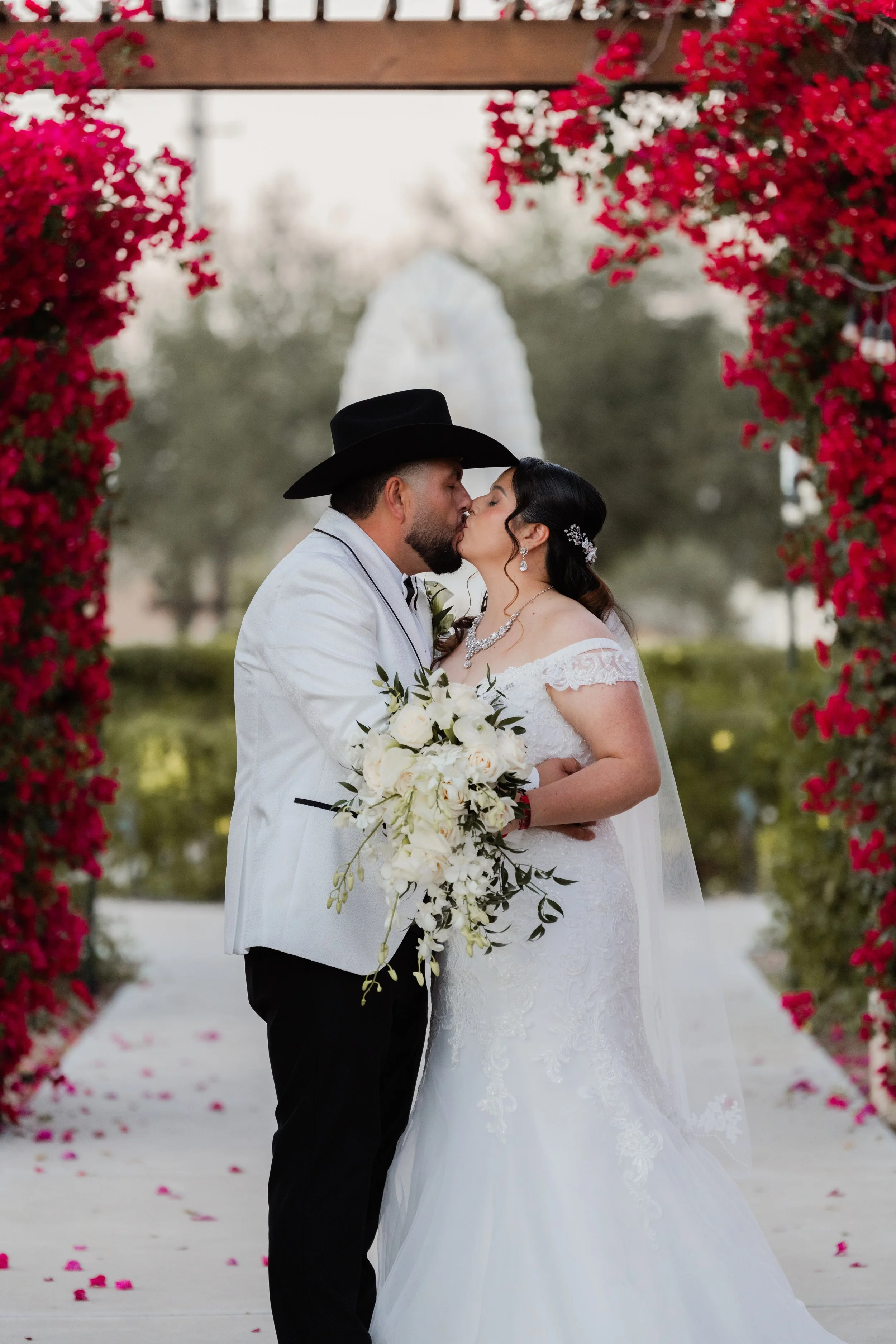 A newlywed couple sharing a kiss during their wedding ceremony, standing under a floral archway with pink flowers, with the White House visible in the background.
