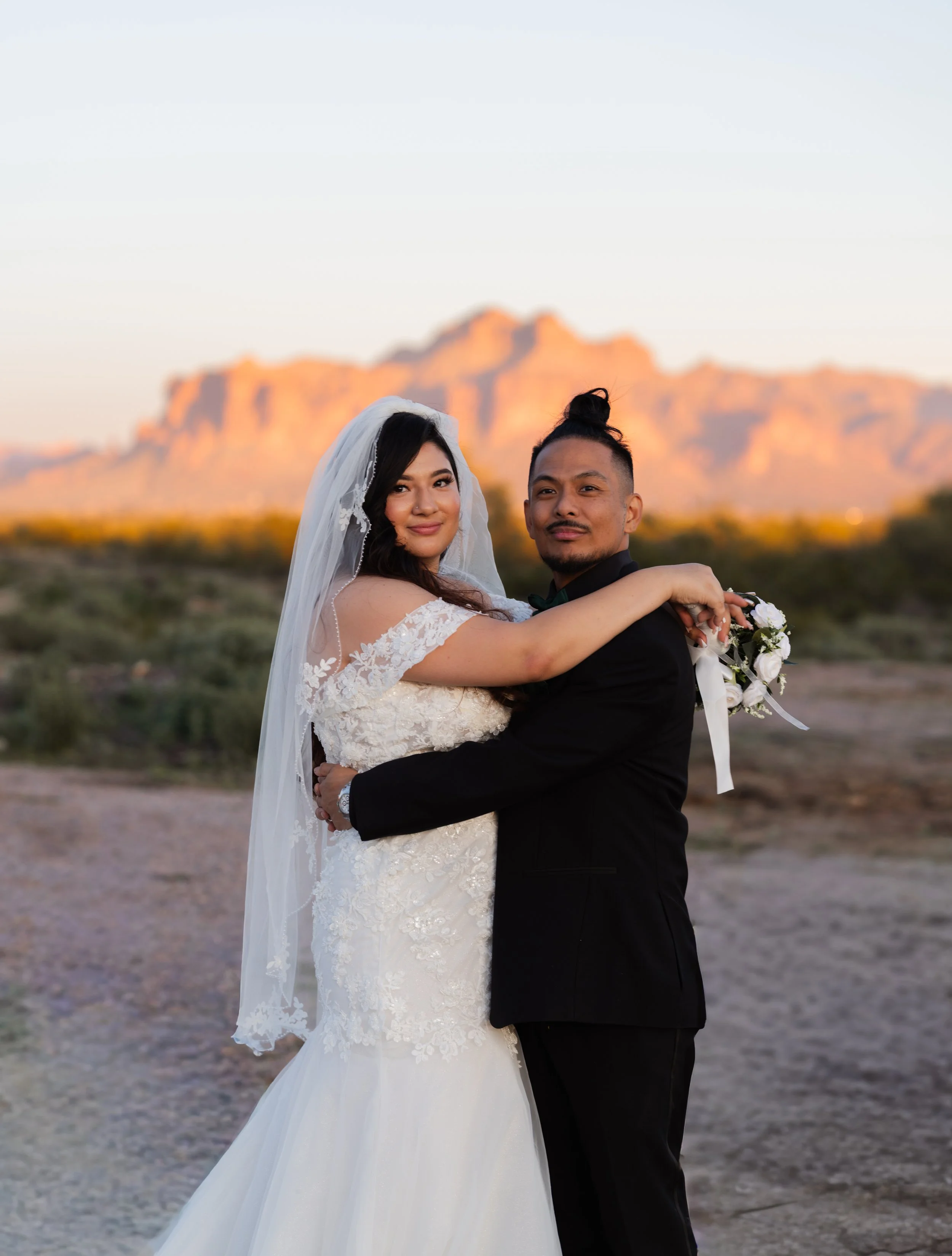 A bride and groom embrace outdoors with mountains in the background, during sunset.