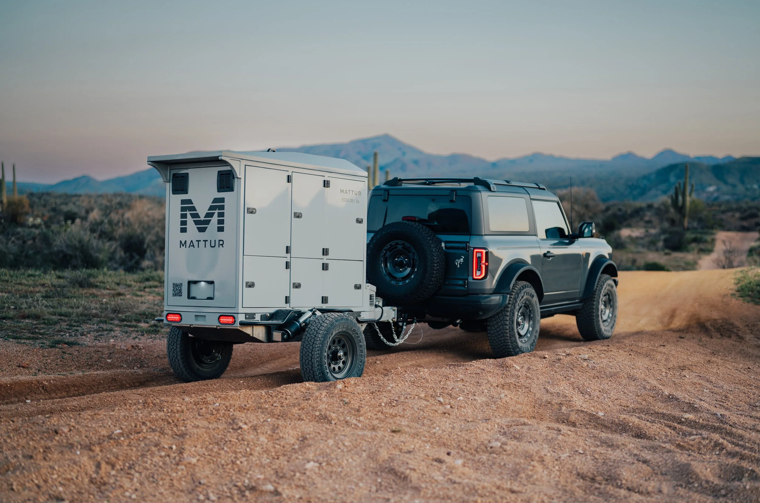 A silver off-road vehicle towing a trailer through a desert landscape with mountains and cacti in the background during sunset.
