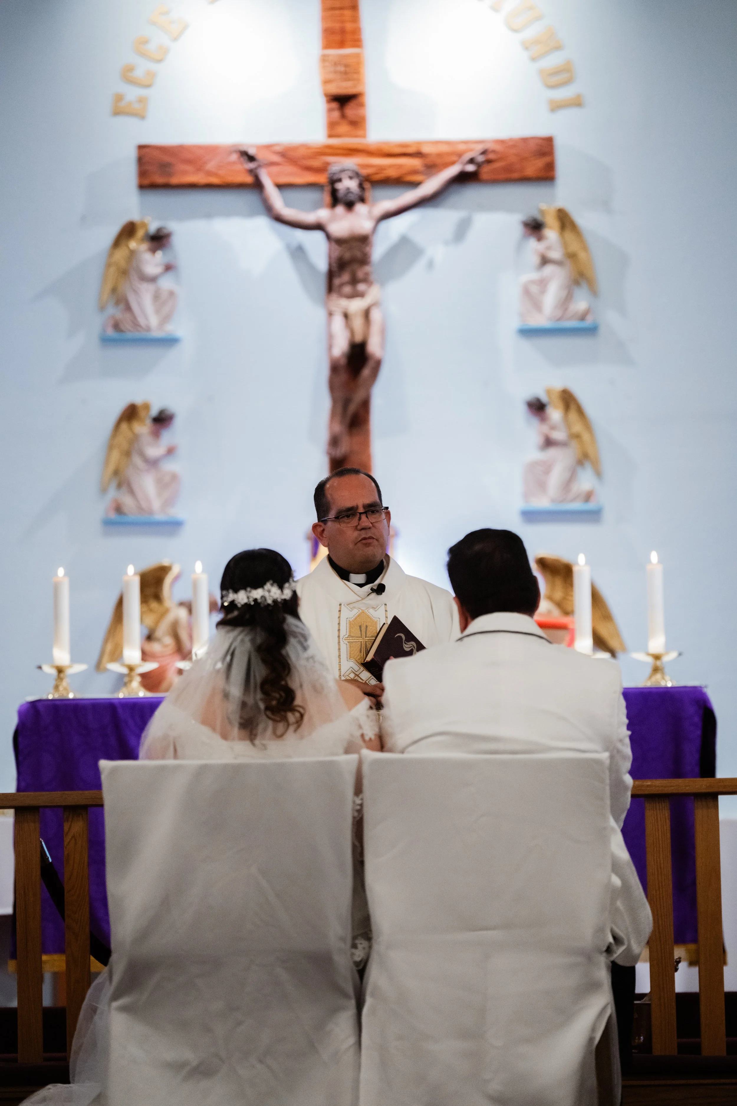 A wedding ceremony held in a church with a priest officiating. The bride and groom are seated facing the priest, with a backdrop of a large crucifix on the wall and angel statues on either side. Candles are lit on the altar.