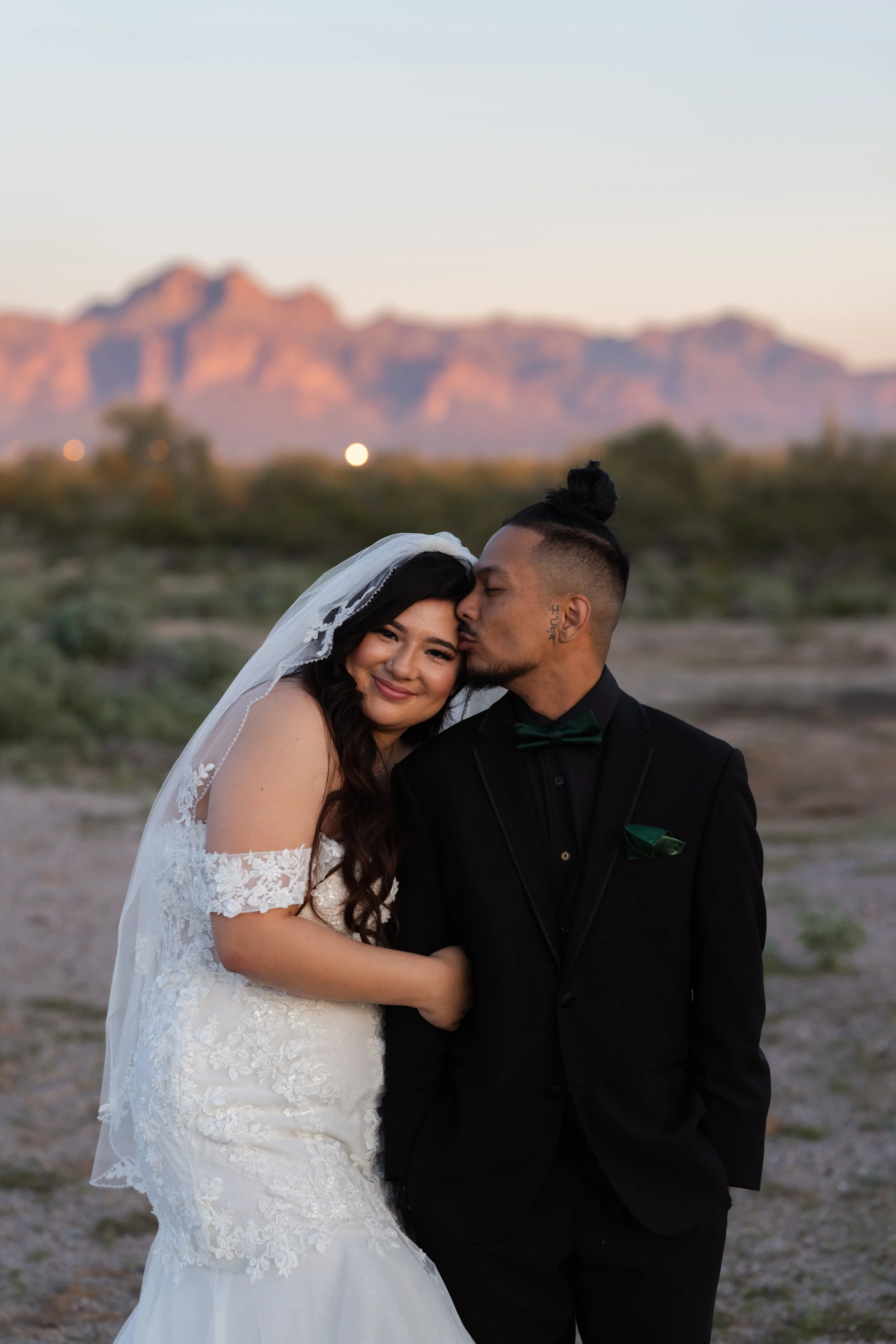 A newlywed couple in wedding attire sharing a tender moment outdoors with mountain scenery in the background.