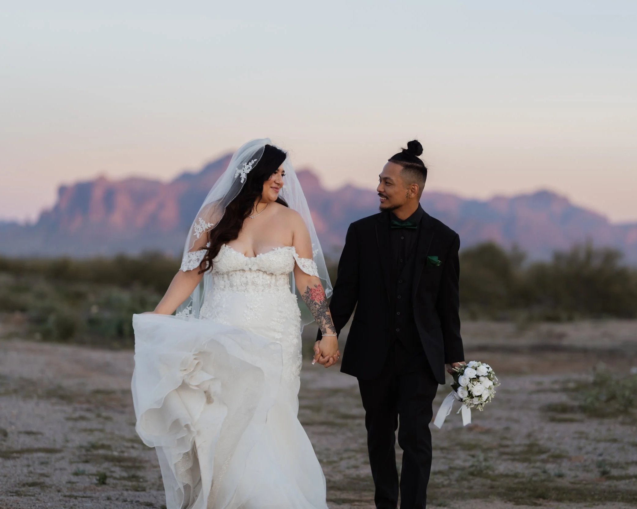 A bride and groom walking hand in hand outdoors during sunset, with mountains in the background. The bride wears a white wedding dress with lace detailing and a veil, and the groom wears a black suit with a green bow tie, holding a bouquet of white flowers.