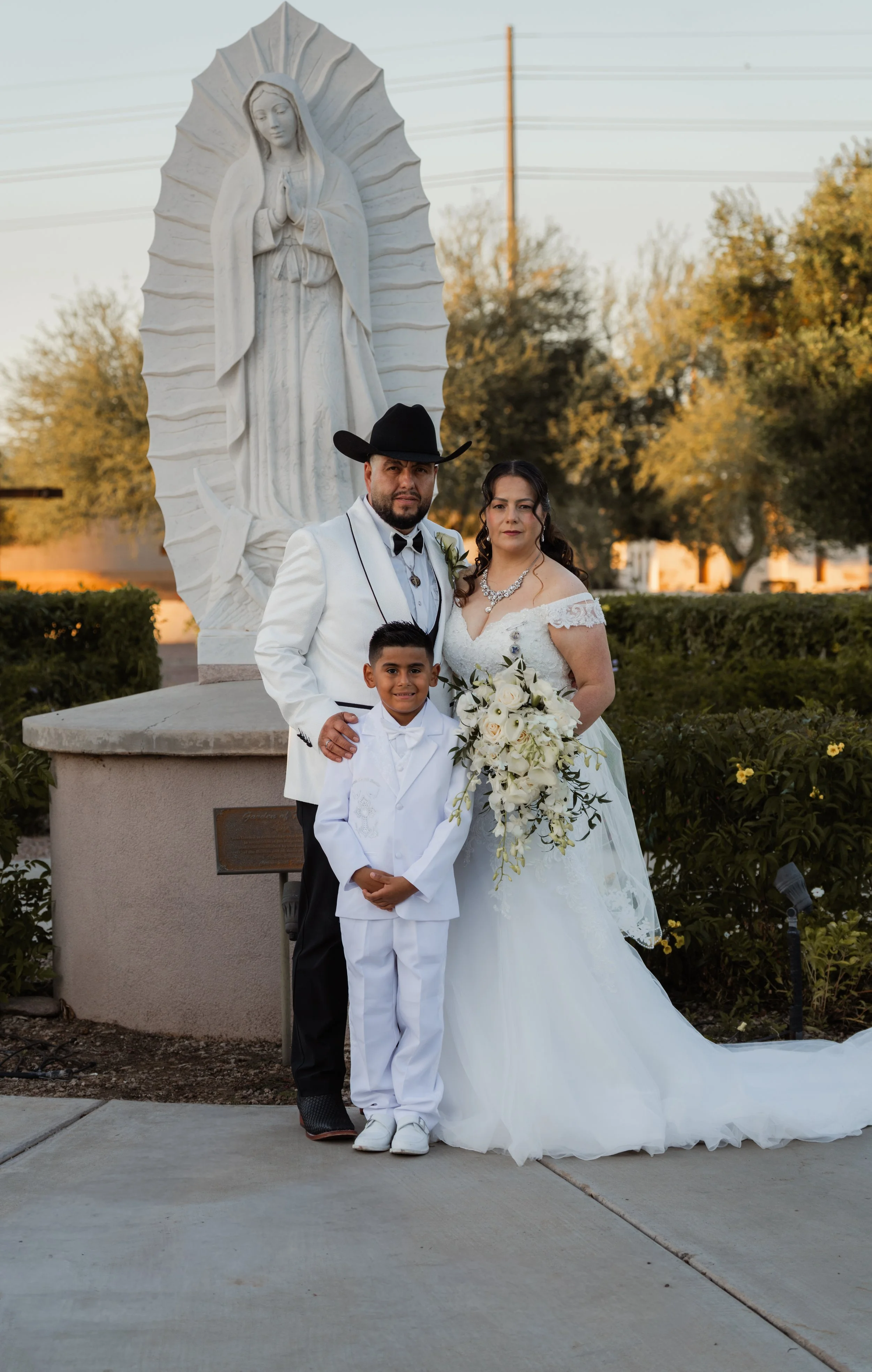 A wedding photo of a couple and a young boy standing outdoors in front of a large religious statue of the Virgin Mary, with trees and power lines in the background. The bride is in a white wedding dress holding a bouquet of white flowers, and the groom is in a white suit with a black cowboy hat. The young boy is dressed in an all-white suit.