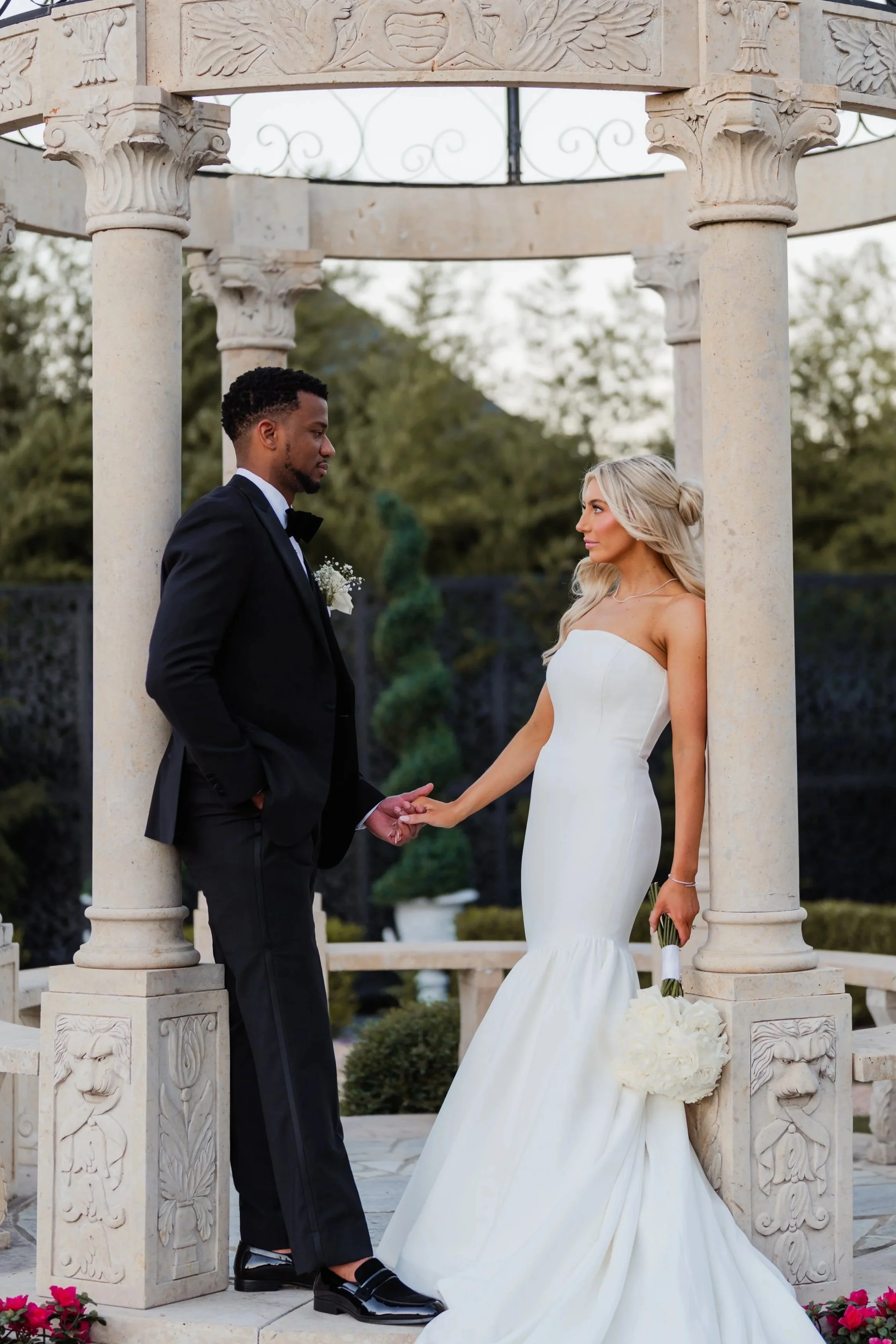 A bride and groom standing underneath a decorative stone gazebo, holding hands and looking at each other during their wedding ceremony. The bride is wearing a strapless white wedding gown and holding a bouquet of flowers, while the groom is in a black tuxedo with a white boutonniere. The setting is outdoors with trees in the background.