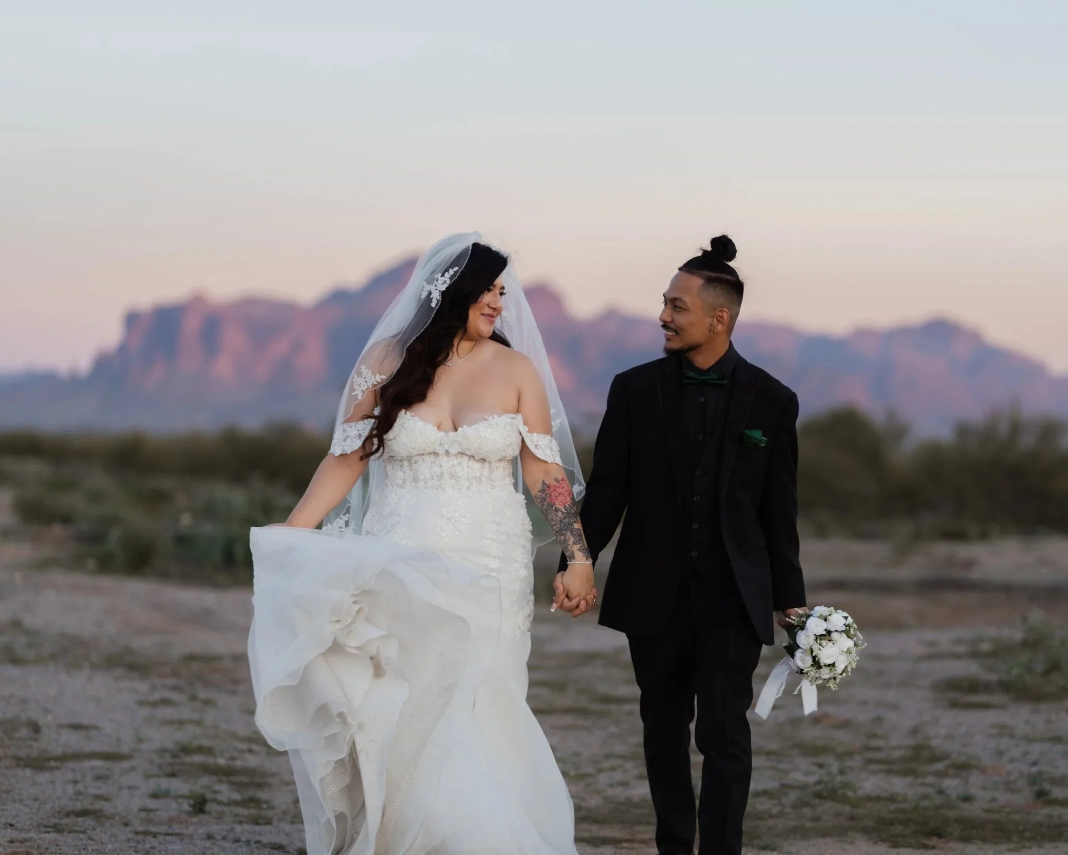 A bride and groom holding hands and walking outdoors at sunset with mountains in the background, the bride in a white wedding dress with a veil and the groom in a black suit holding a bouquet of white flowers.