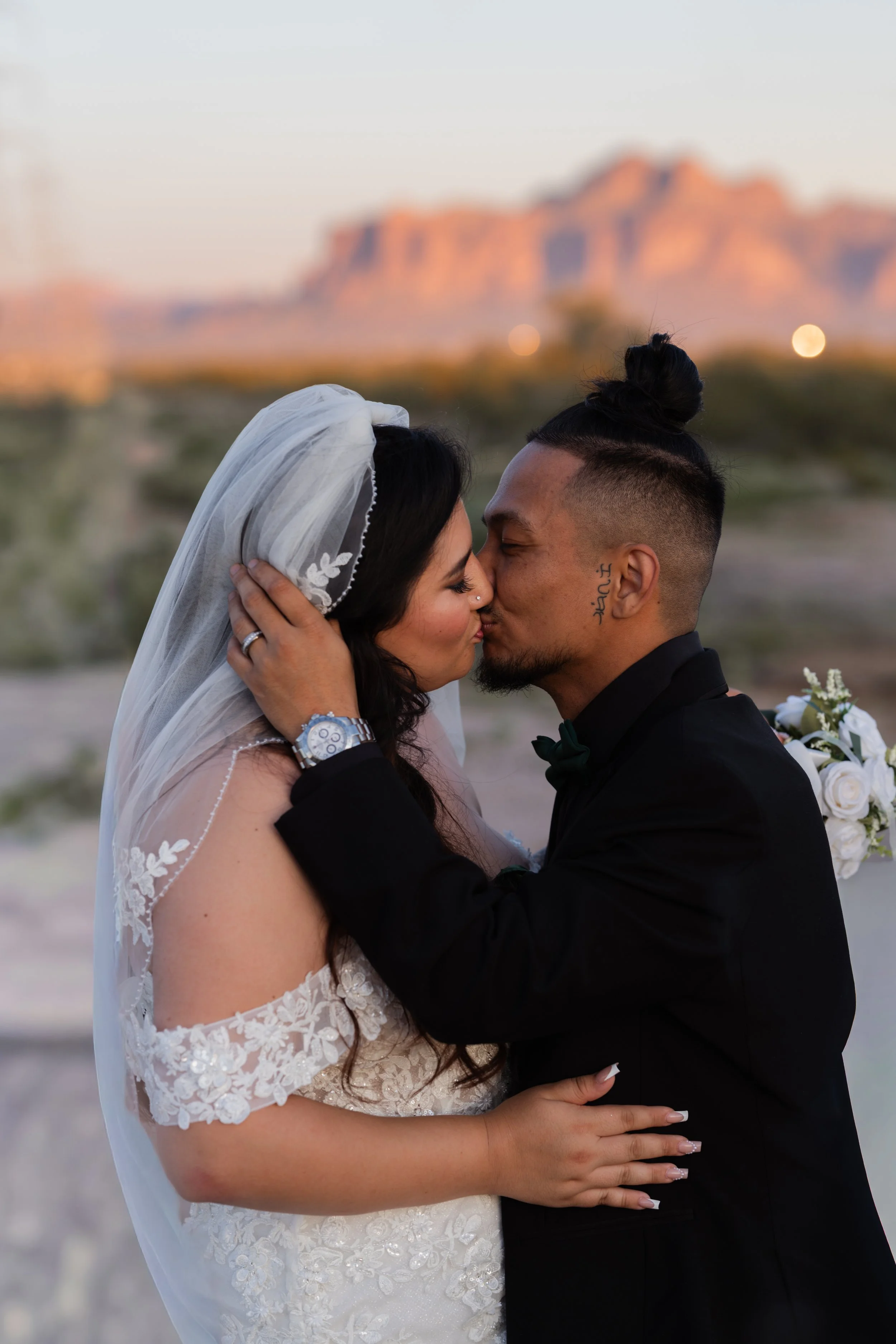 A newlywed couple kissing outdoors at sunset with mountains in the background.