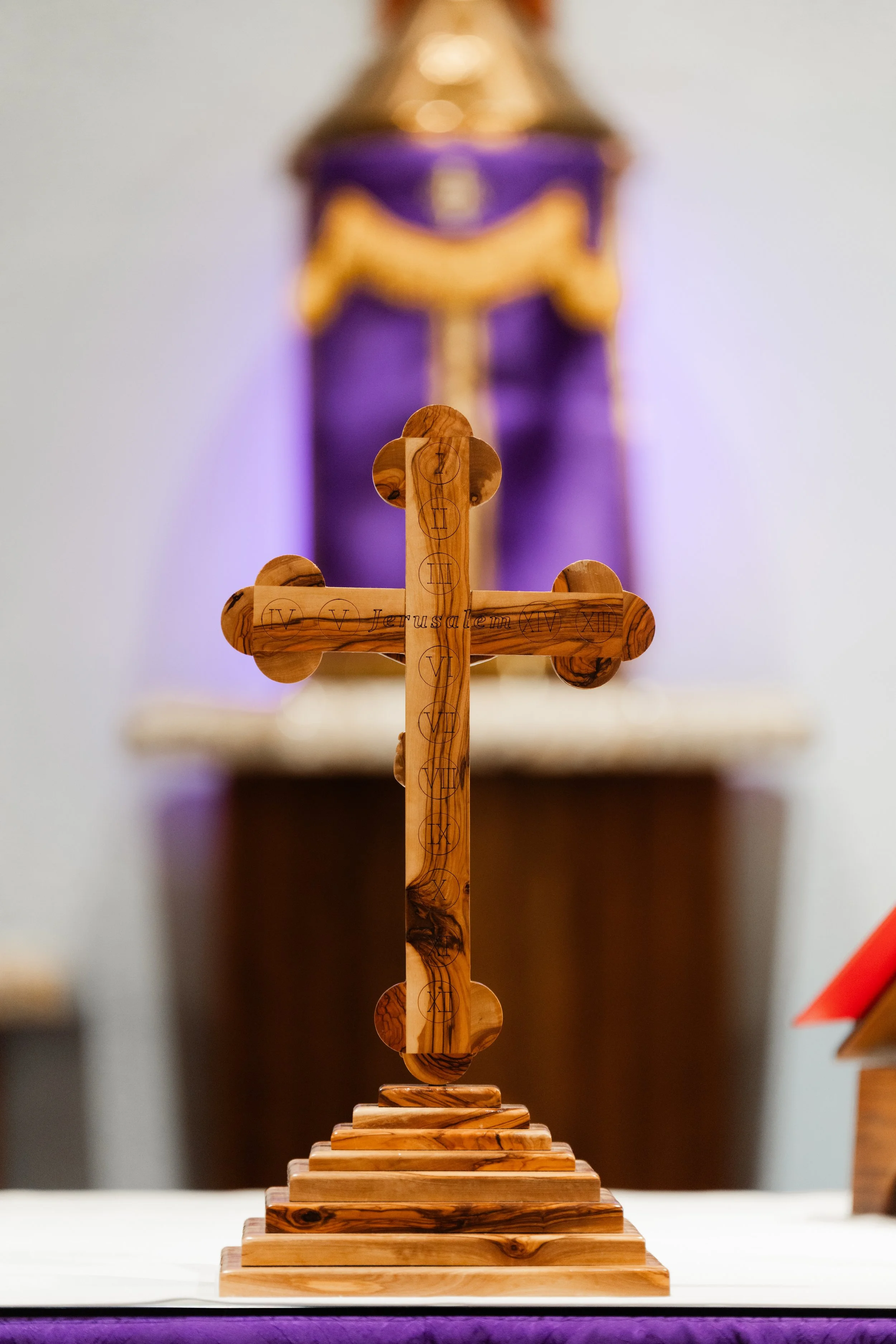 Wooden cross with Roman numerals and the inscription "Jerusalem" on it, placed on a stepped wooden base, with a blurred religious altar or statue in the background.