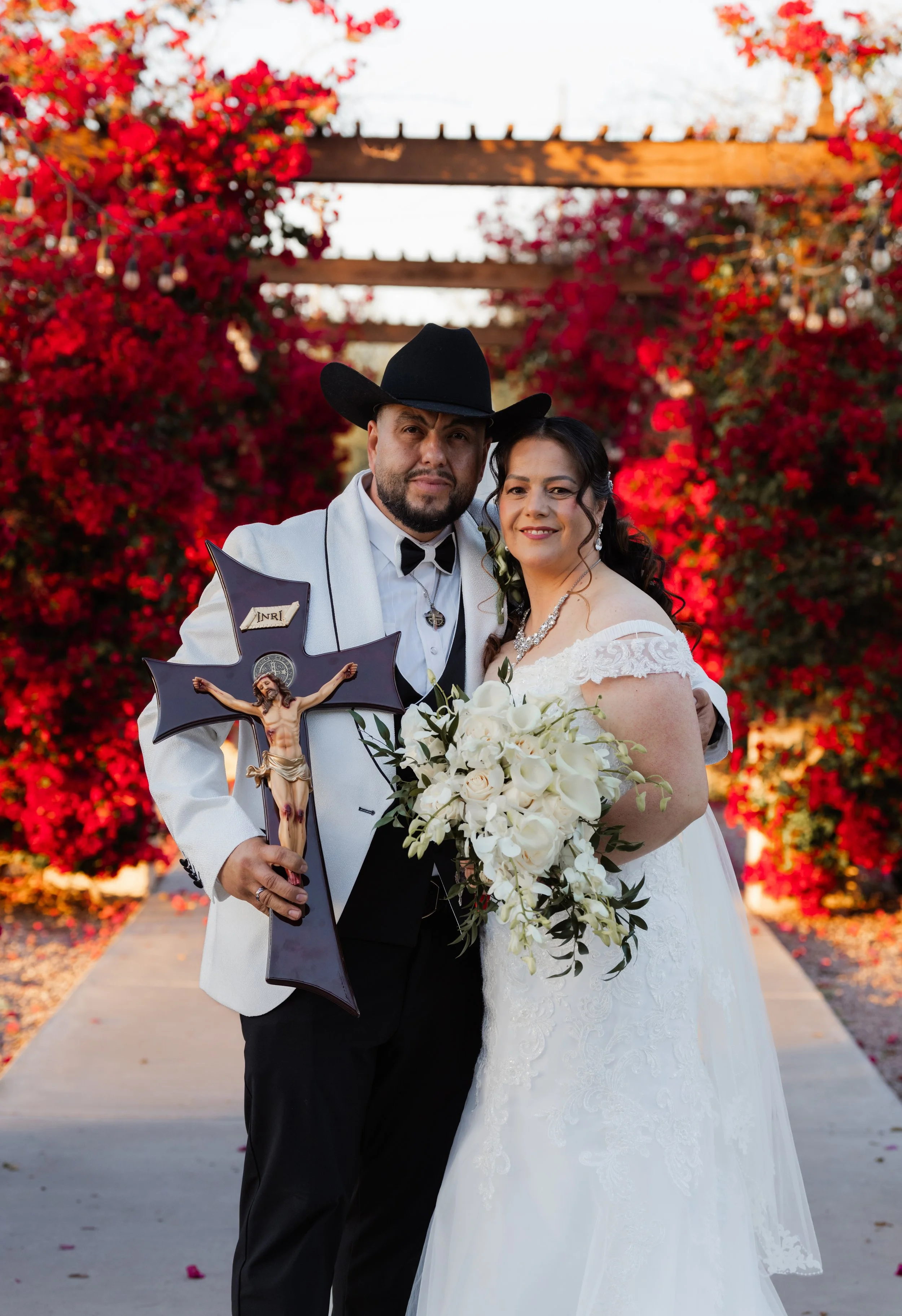 A couple in wedding attire posing outdoors in front of vibrant red flowering bushes, with the groom holding a crucifix and the bride holding a bouquet of white flowers.