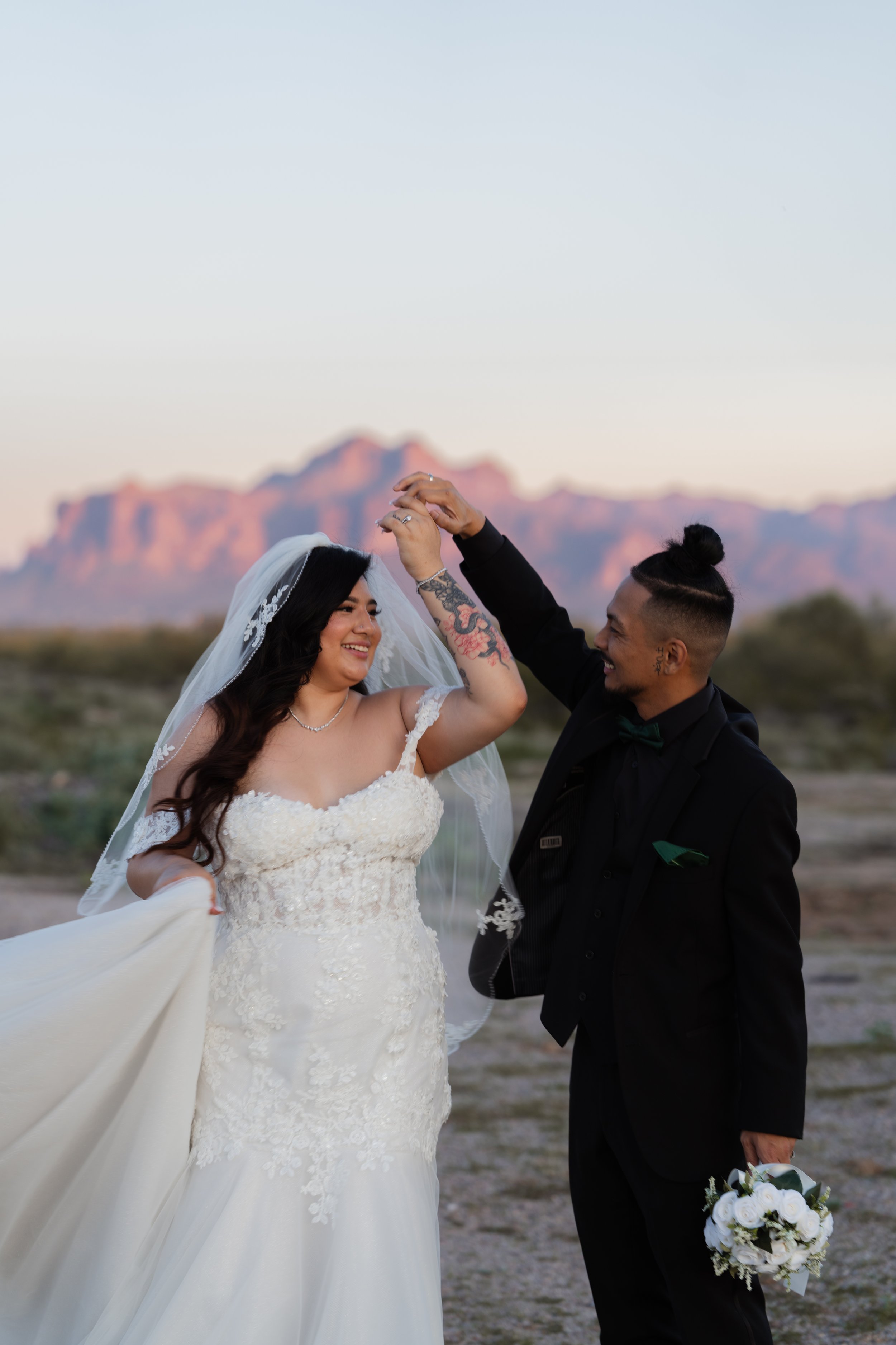 Bride and groom dancing outdoors at sunset, with mountains in the background, the bride in a white wedding gown and veil, and the groom in a black suit holding a bouquet.