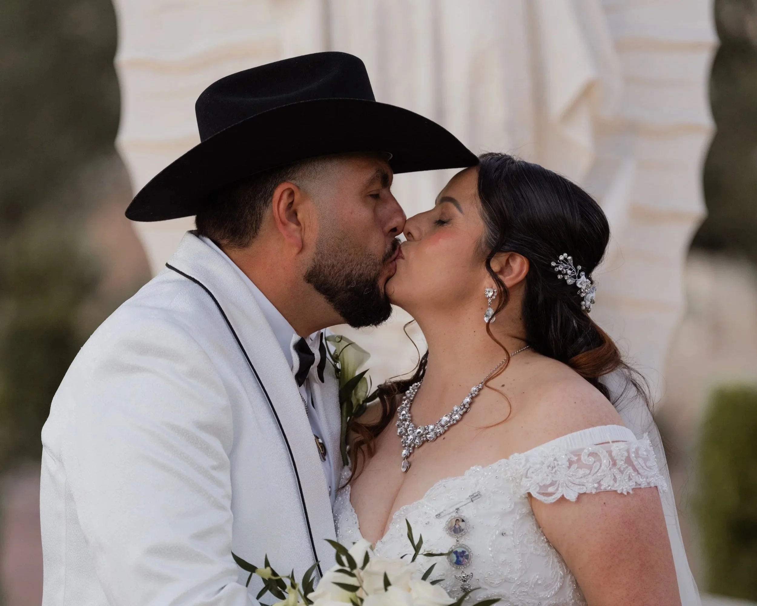 Bride and groom kissing at their wedding, groom wearing a black cowboy hat and white suit with black trim, bride in a white lace off-shoulder dress with ornate jewelry, holding a bouquet of white flowers.