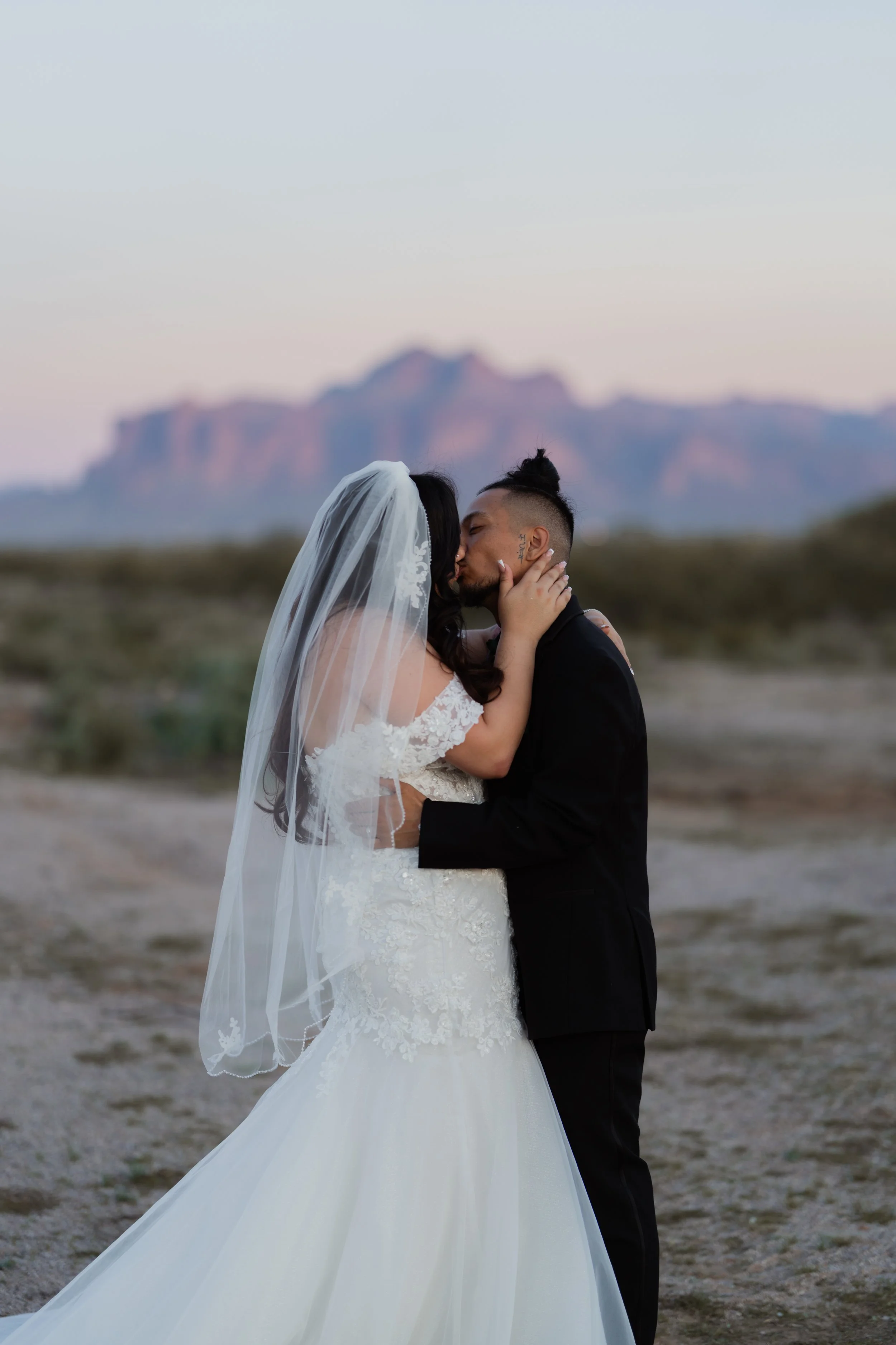 A couple in wedding attire kissing outdoors with mountains in the background.