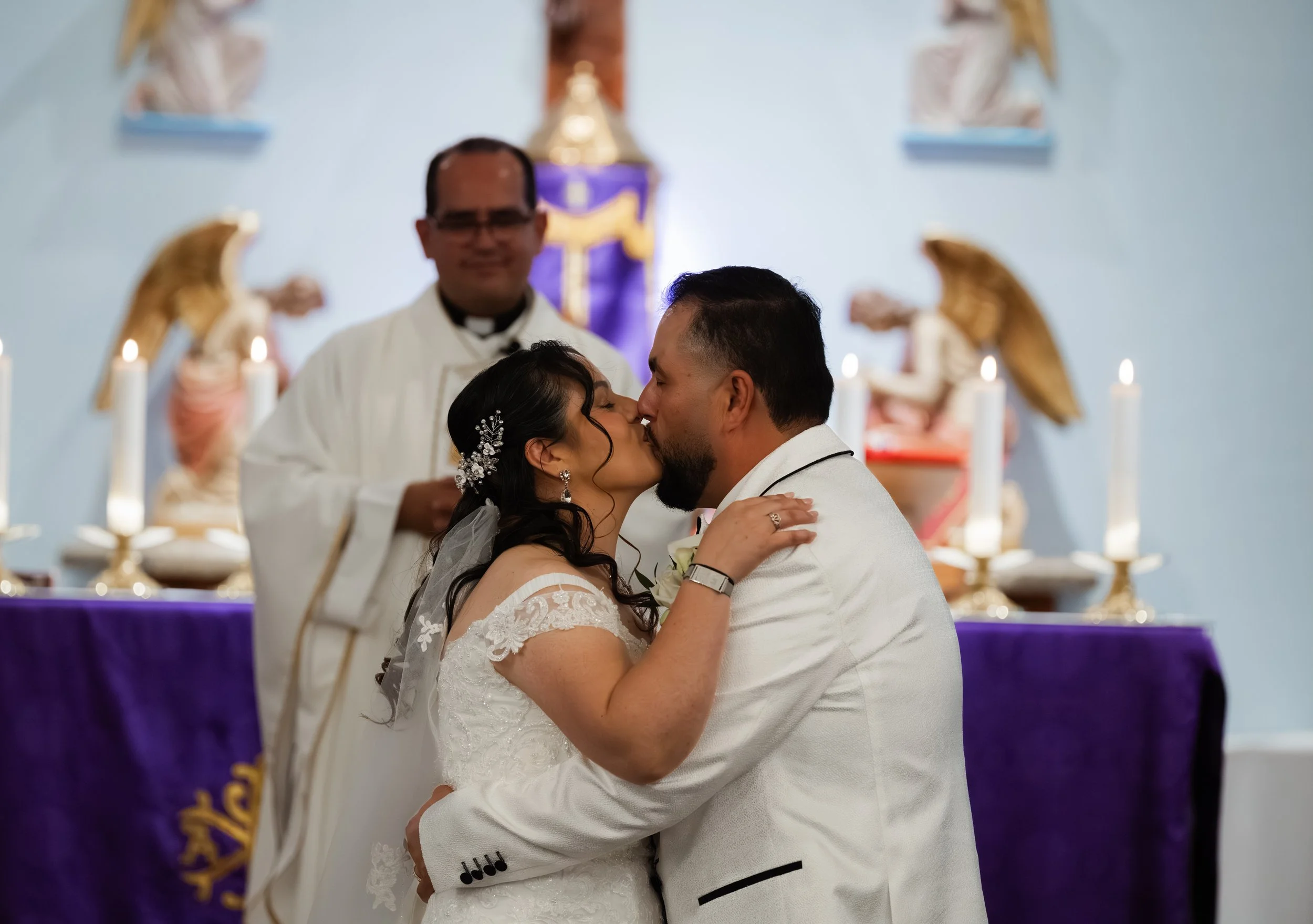A bride and groom kissing during their wedding ceremony standing in front of a priest inside a church, with religious figures and candles in the background.