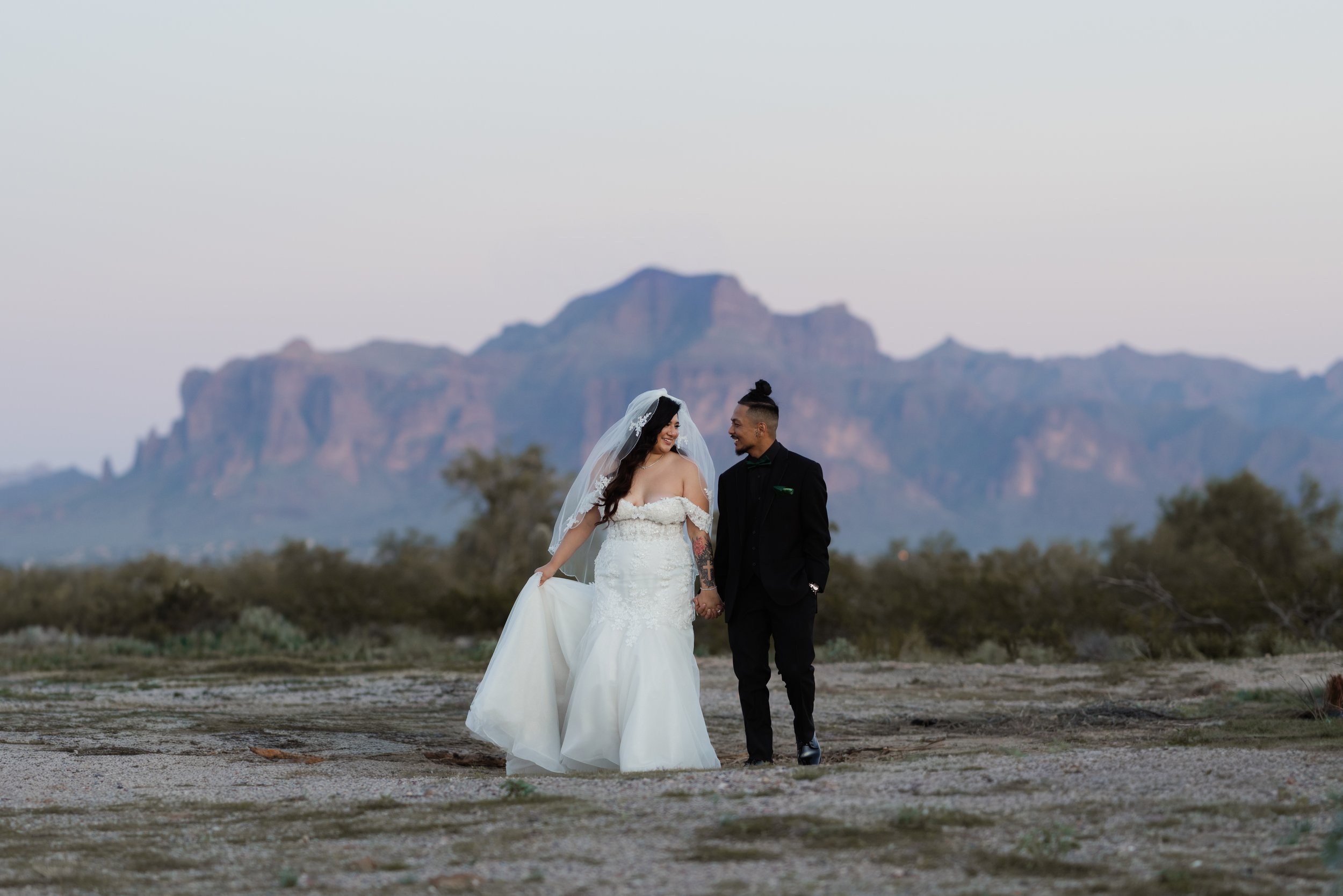 A bride and groom walk hand in hand in a desert landscape with mountains in the background, dressed in their wedding attire.